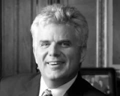 Black and white photo of a smiling man with light-colored hair, wearing a suit and tie, standing in front of wooden paneling.