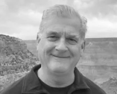 Black and white photo of a smiling man with short, light-colored hair, wearing a dark jacket, outdoors near a canyon or cliff.