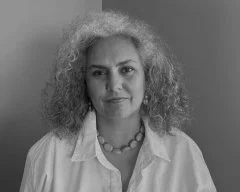 A woman with curly hair wearing a necklace and a light-colored blouse, looking at the camera in a grayscale photo.