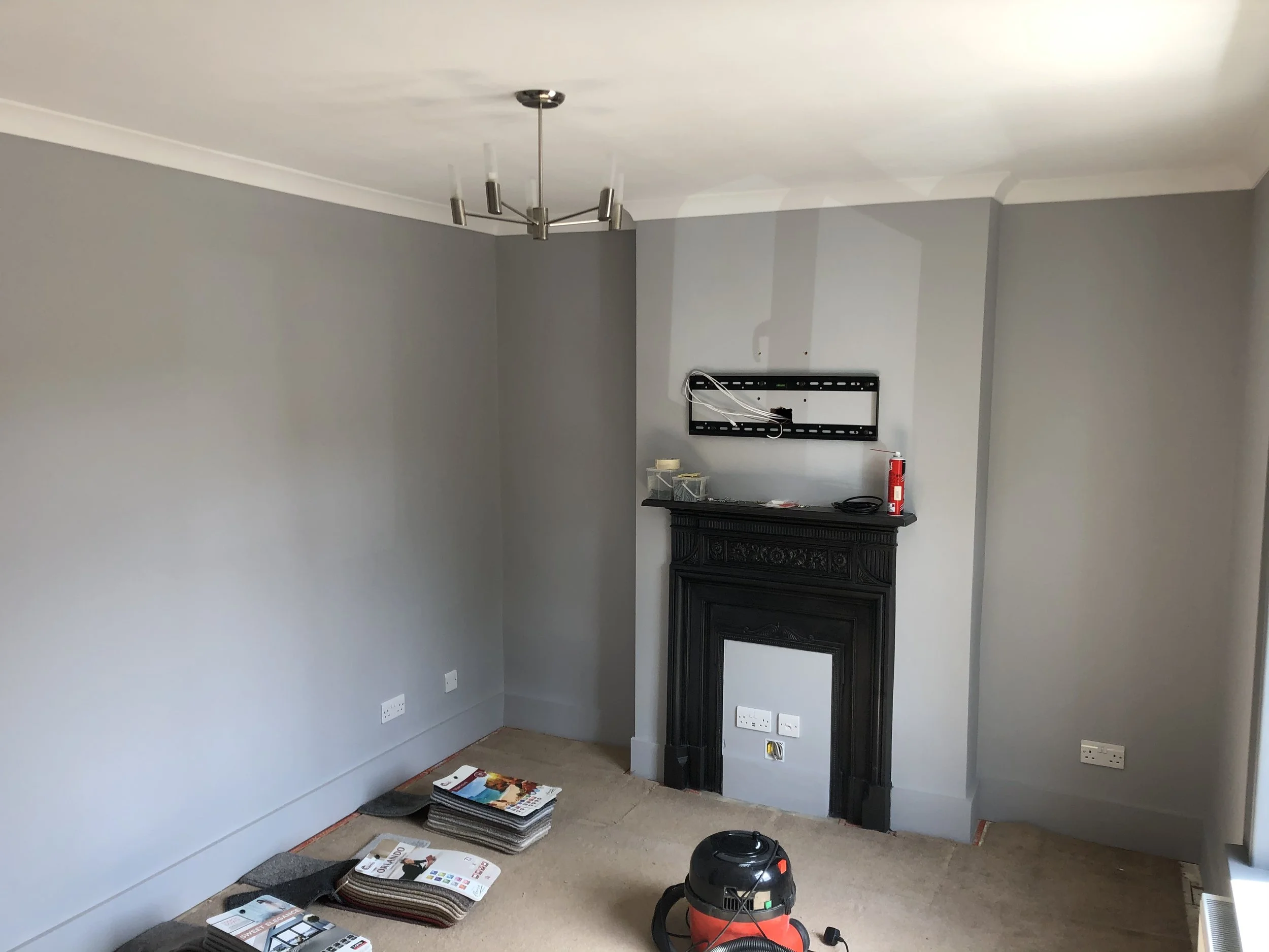Living room with grey walls, black fireplace mantel, ceiling light fixture, and various items on the floor and mantel, including a vacuum cleaner, magazines, and electronic accessories.