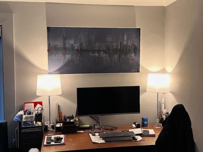 A well-organized home office desk with two table lamps on either side, a widescreen monitor, various office supplies, and a black office chair, with a large abstract cityscape painting on the wall behind.