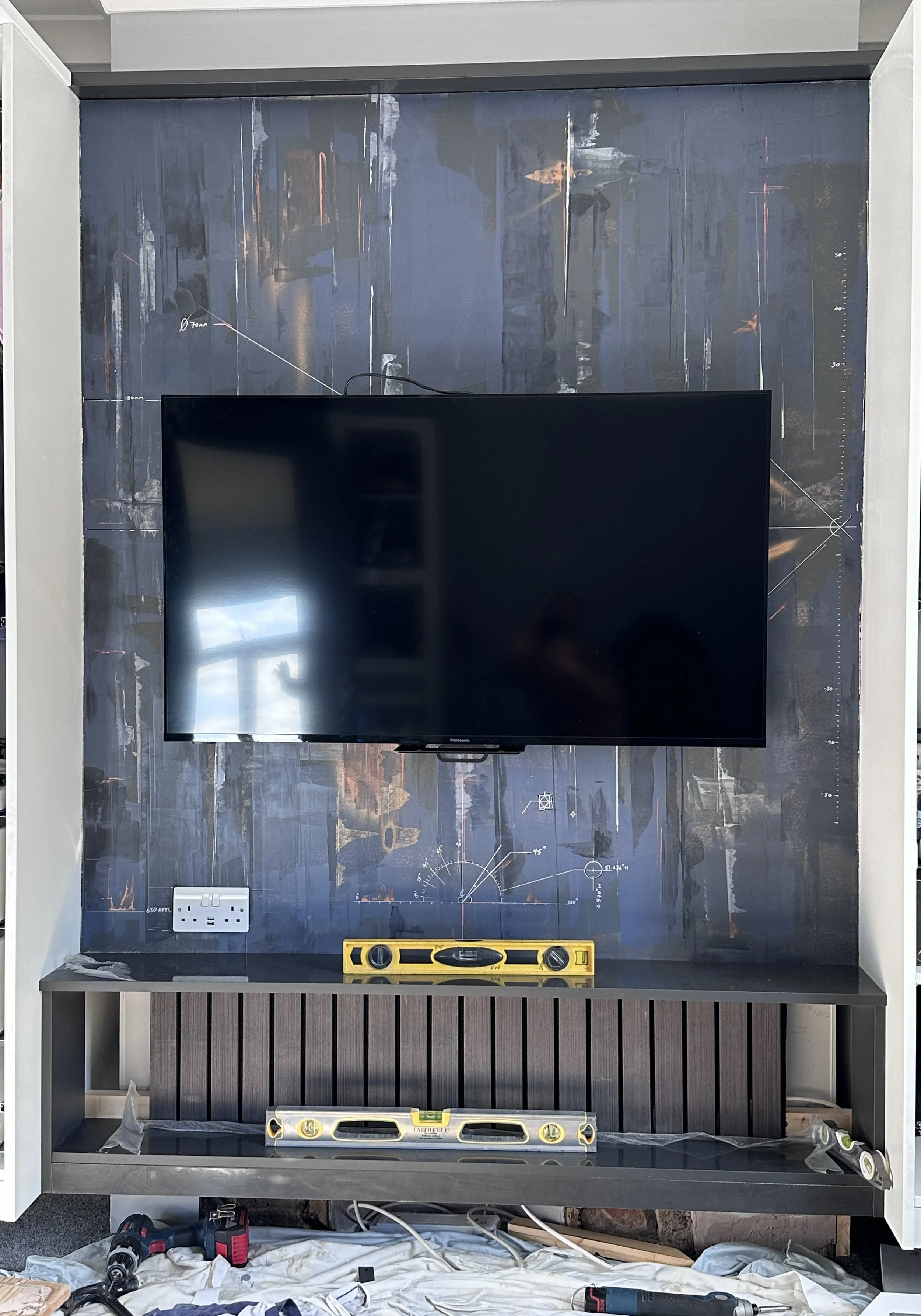 TV wall with a large flat-screen TV mounted on the wall, surrounded by construction tools including levels, a drill, and a pipe. The background features a dark decorative wall panel with technical drawings and measurements.
