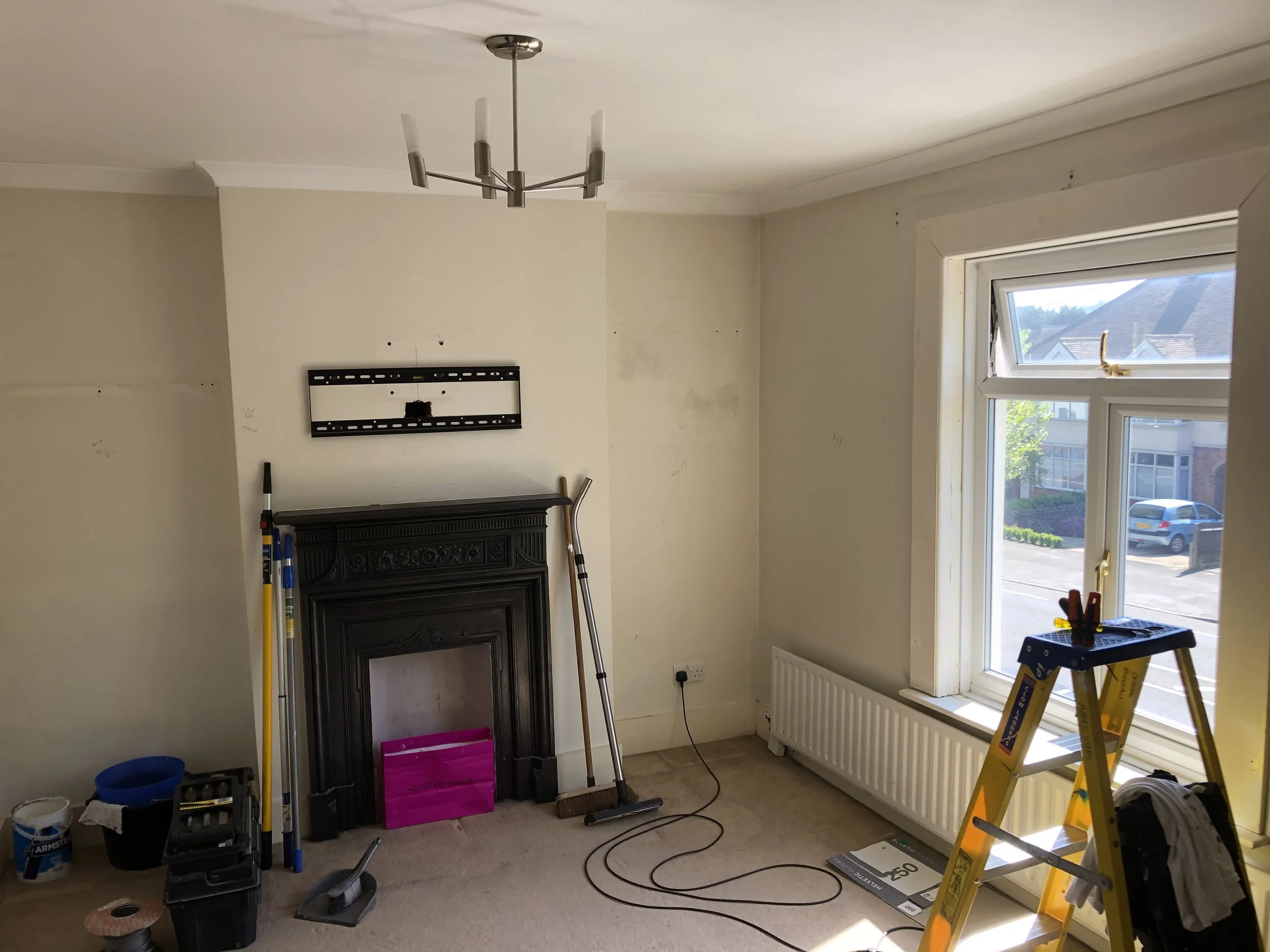 Living room with unfinished walls, a black fireplace, a yellow step ladder, tools, and a large window showing a residential street outside.