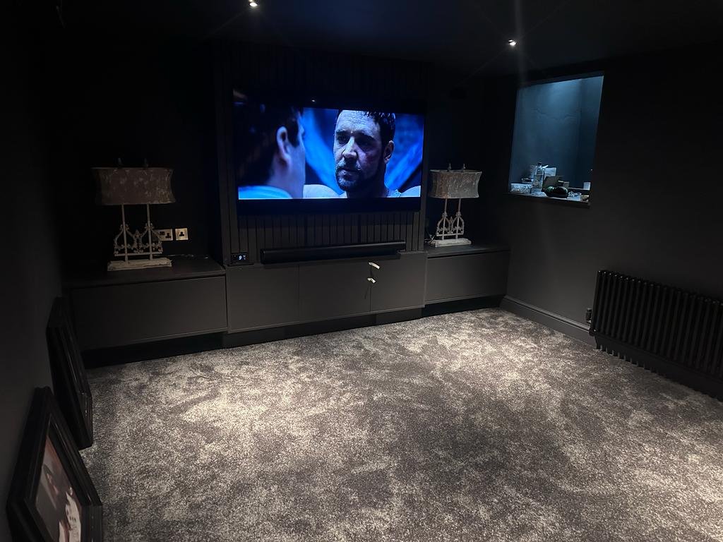 A dark home theater room with a large flat-screen TV showing a movie scene with two men in conversation, one with facial hair. The room has dark walls, two lamps with ornate lampshades on either side of a low sideboard, and a window or opening with a view of another room. The floor is covered with a textured grey carpet, and there is a radiator on the right wall.