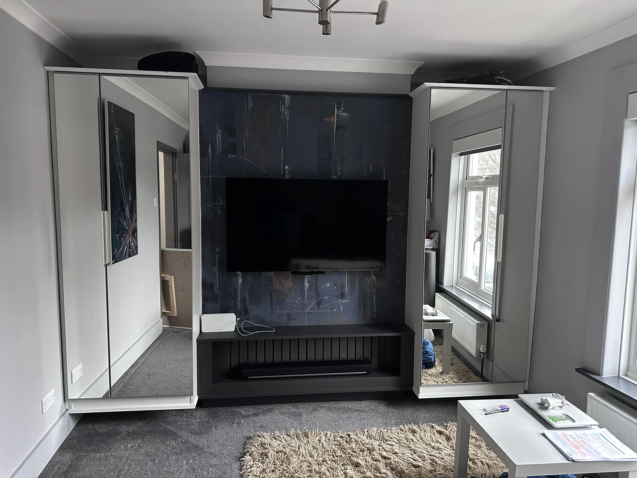 Living room with a wall-mounted television and mirrored wardrobe cabinets on either side, a beige shag rug, a small white coffee table with magazines and pens, and large windows with white frames.