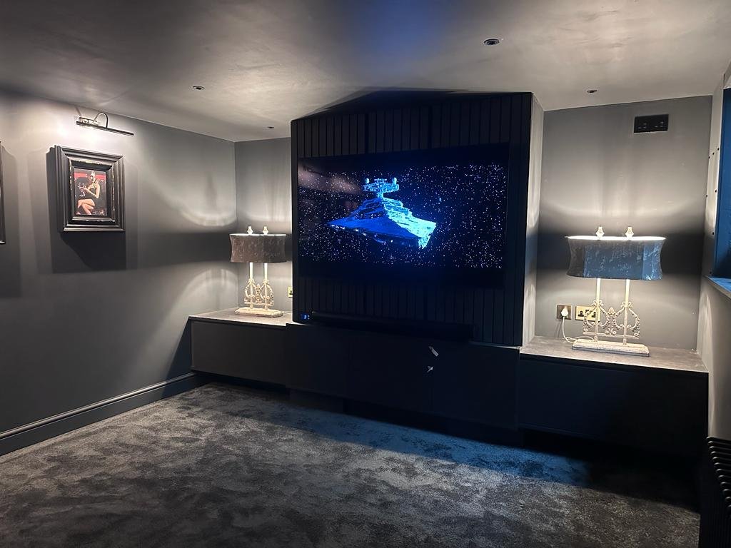 A modern entertainment room with a mounted TV displaying a spaceship, flanked by two table lamps, and decorated with a framed picture on the wall.