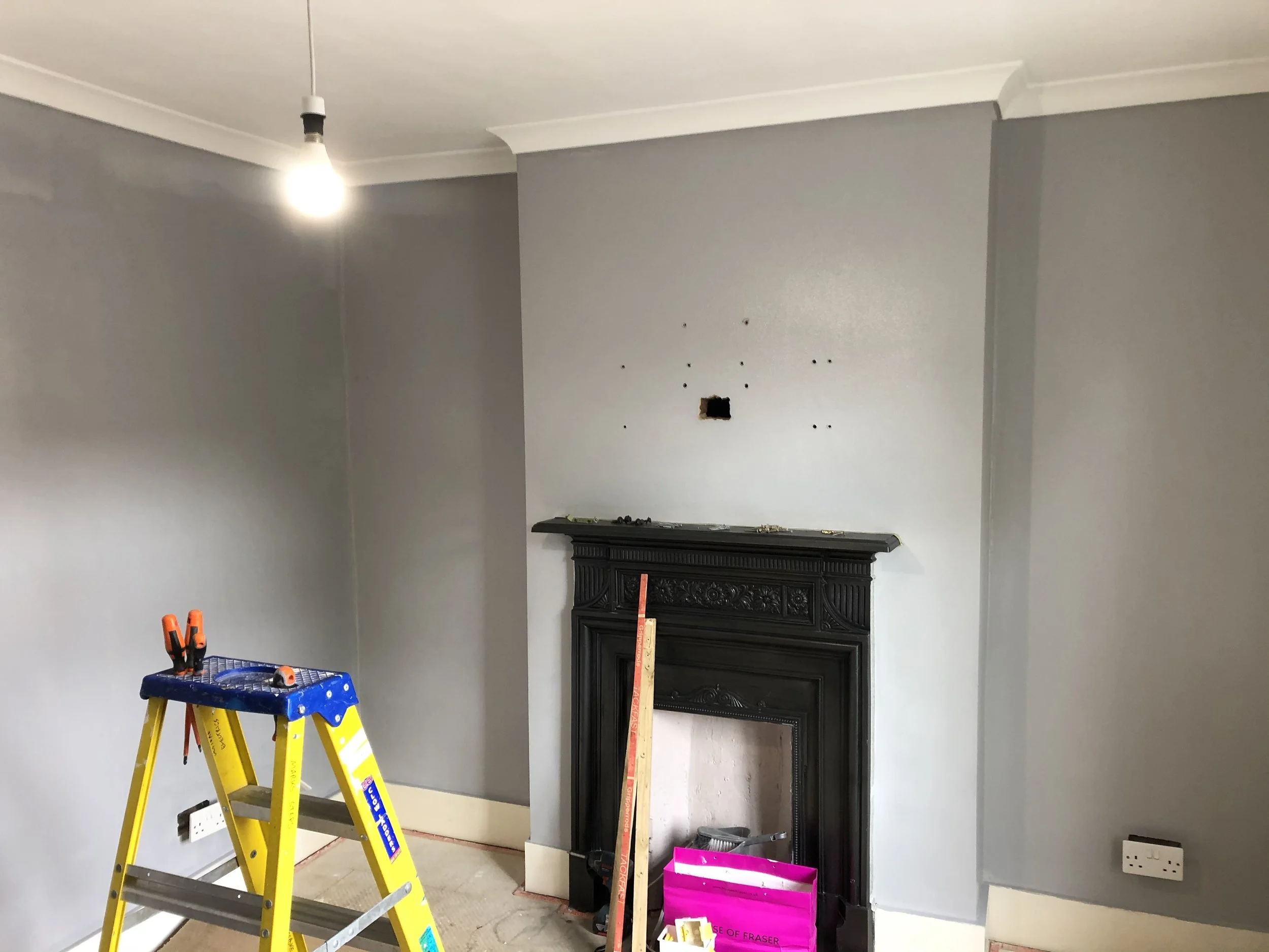 Living room with a gray painted wall, a black fireplace mantel, and tools placed on the floor, including a pink bag and a yellow step ladder near the wall with a light bulb hanging from the ceiling.