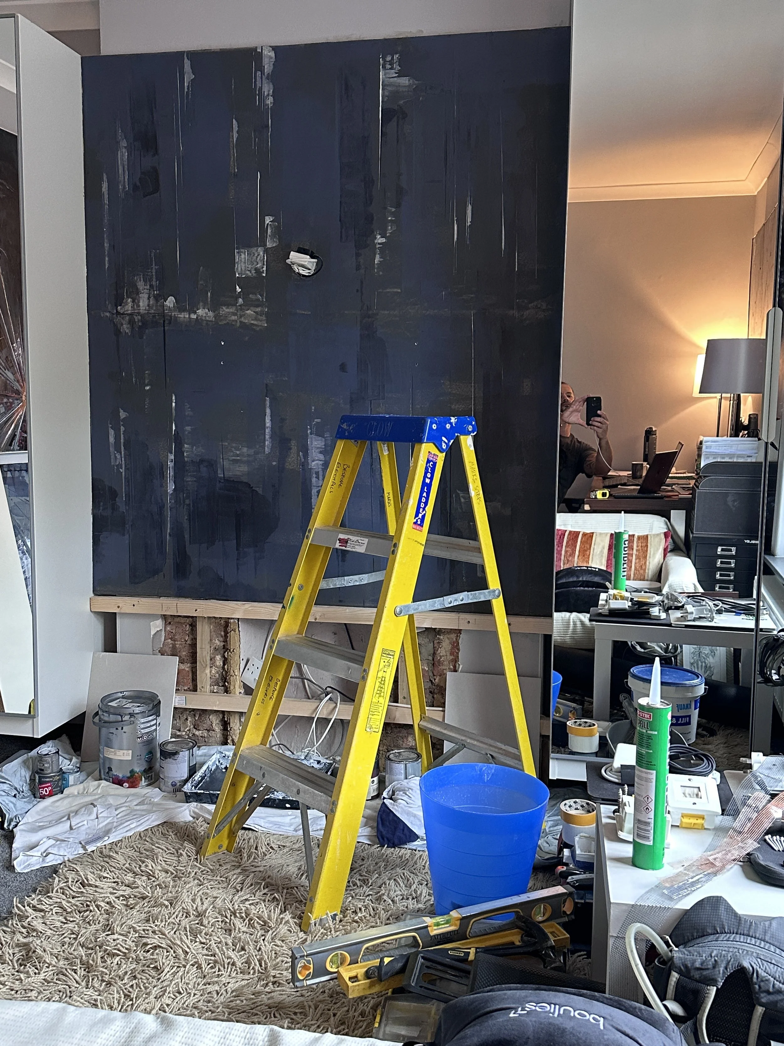 Inside a room under renovation with a yellow ladder in the center, paint cans on the floor, and a black wall with some unpainted areas. A mirror or window reflects a person taking the photo, along with a cluttered desk and lamp in the background.