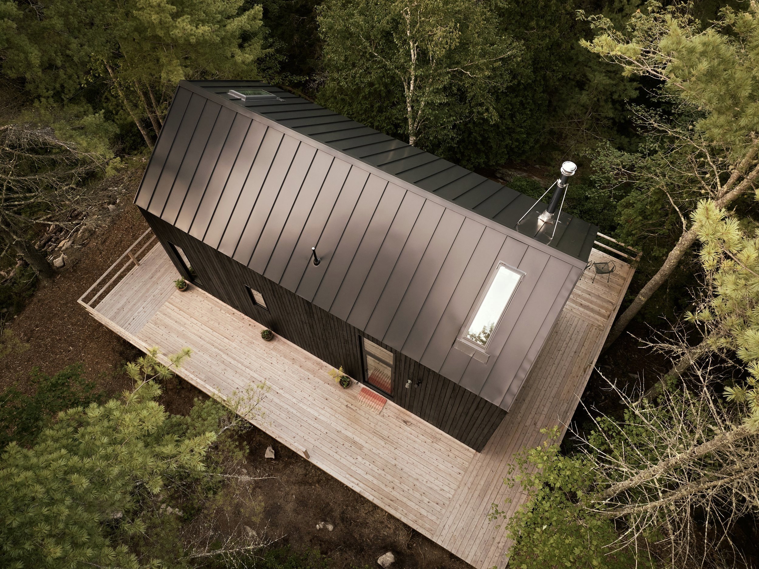 Aerial view of Moss House, a modern home with a metal roof and a wooden deck surrounded by trees and water.