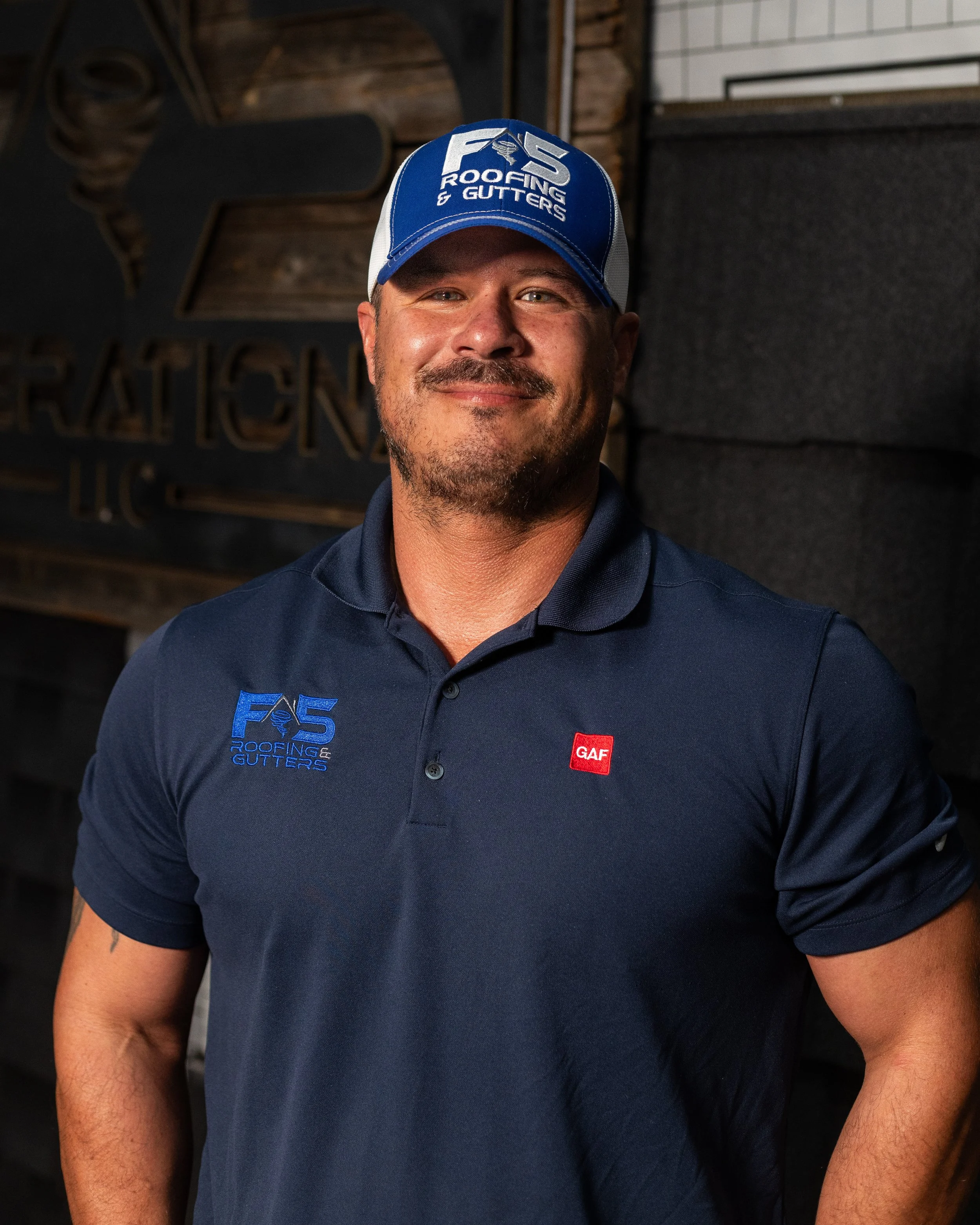 A man wearing a blue cap with 'F5 Roofing & Gutters' logo and a blue polo shirt with the same logo, smiling, in front of a wall with dark and wooden decor.
