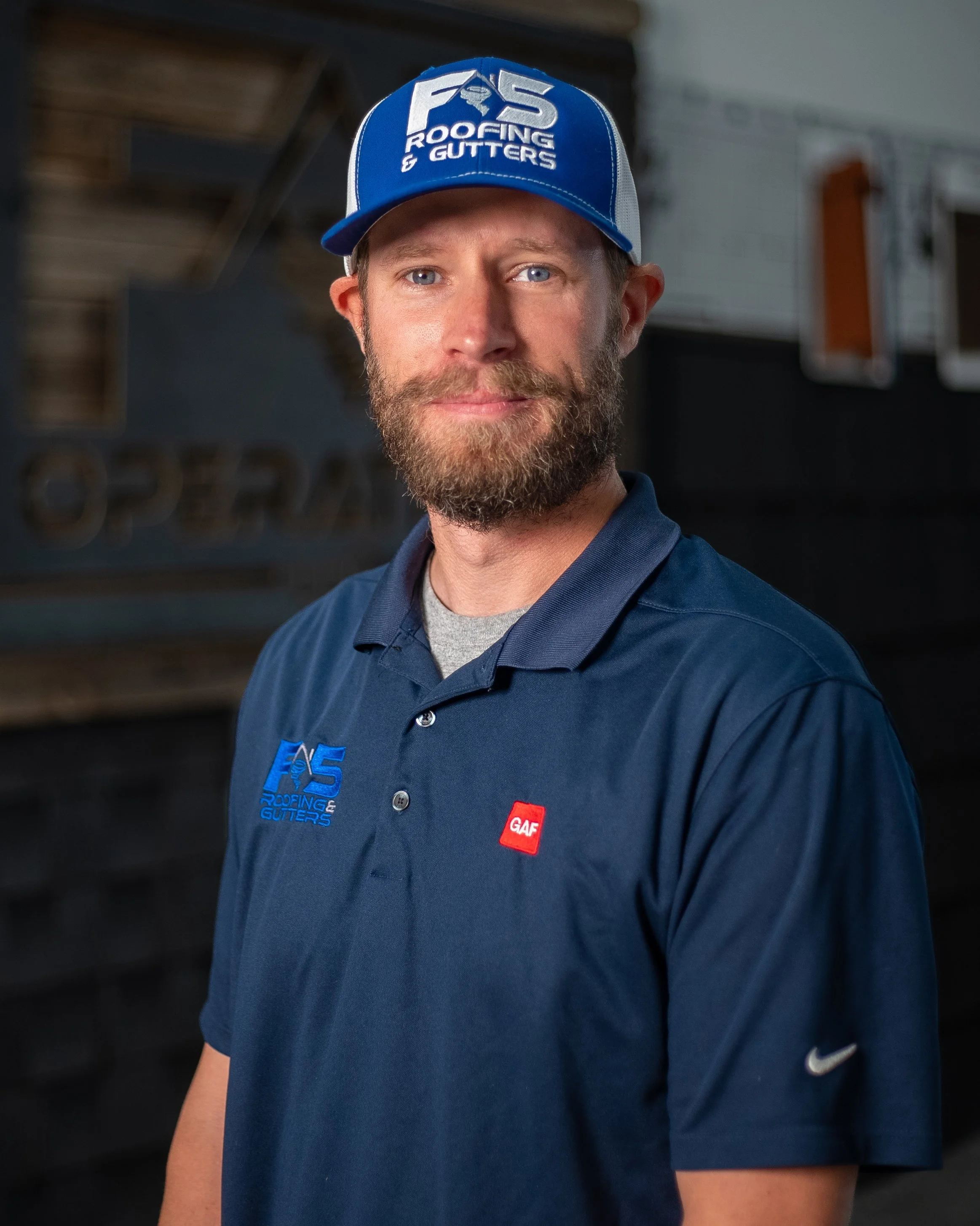 Person with a beard looking into camera with a blue baseball cap on and a navy blue polo.
