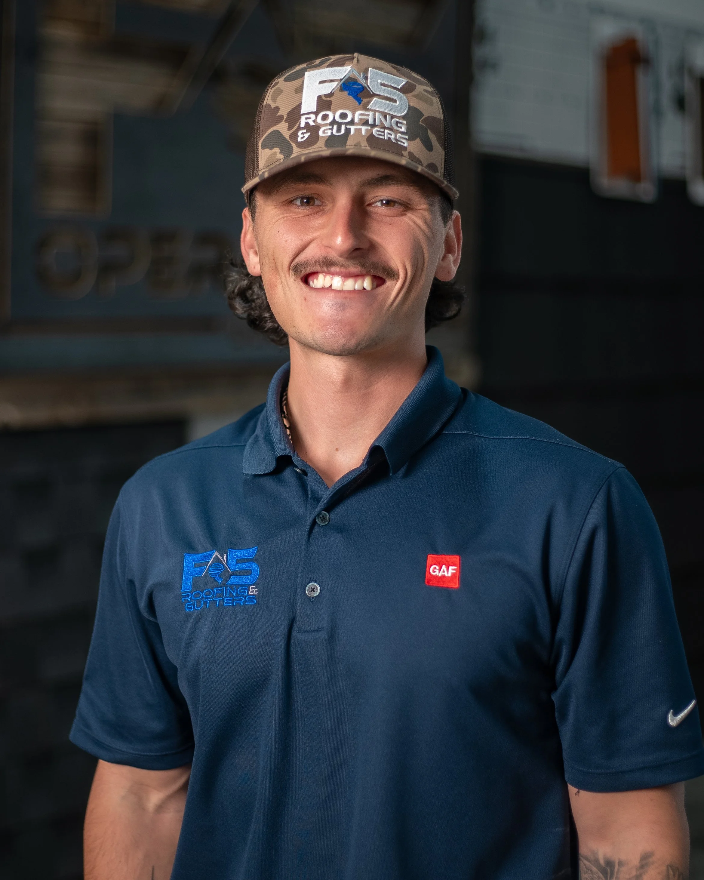 A young man smiling, wearing a blue polo shirt with logos, standing outdoors with a blurred background.