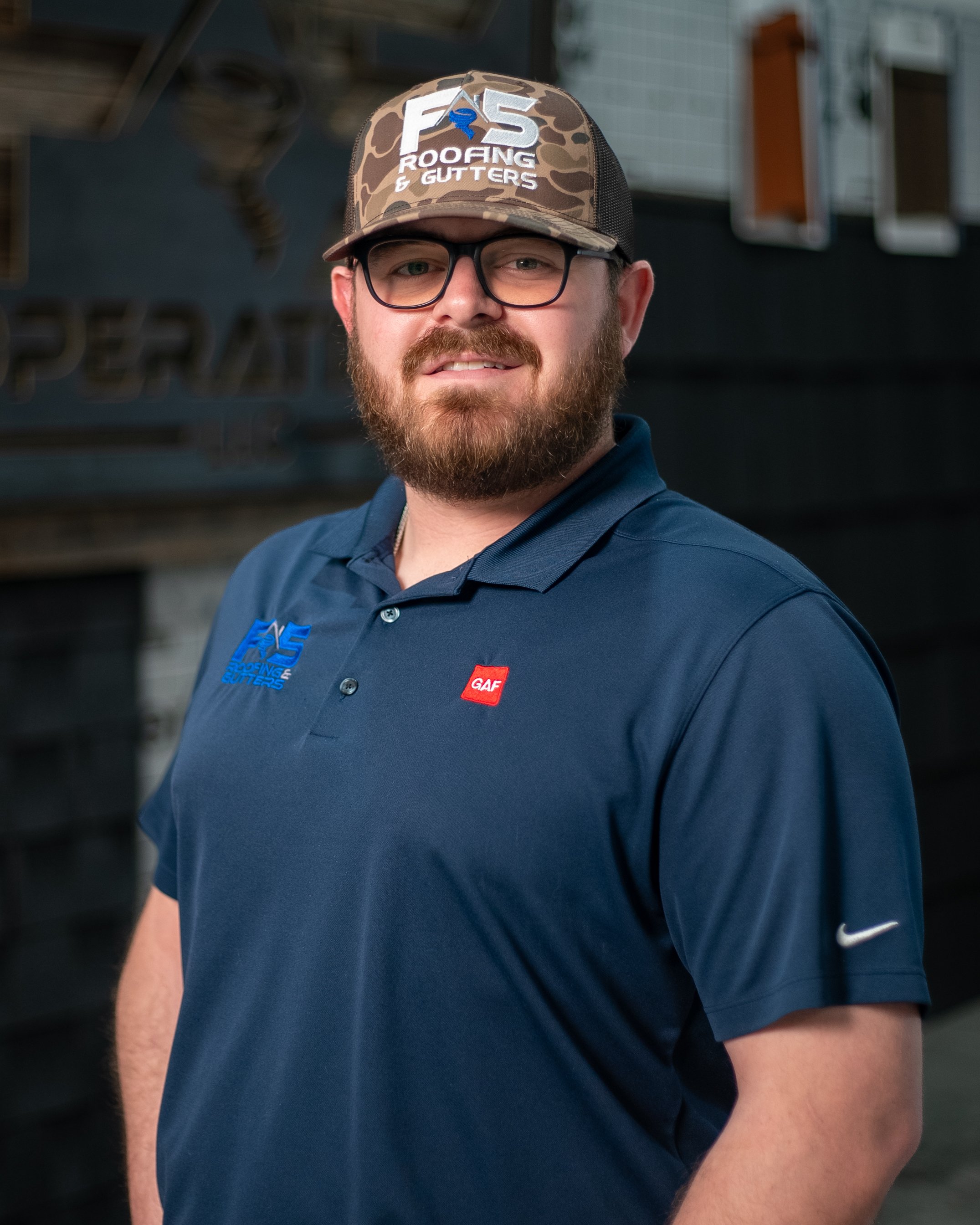 A man with glasses and a beard wearing a black and gray cap with 'F5 Roofing & Gutters' logo, a blue collared shirt with the same logo and an 'Atlas' patch on the sleeve, standing outdoors in front of a glass window.