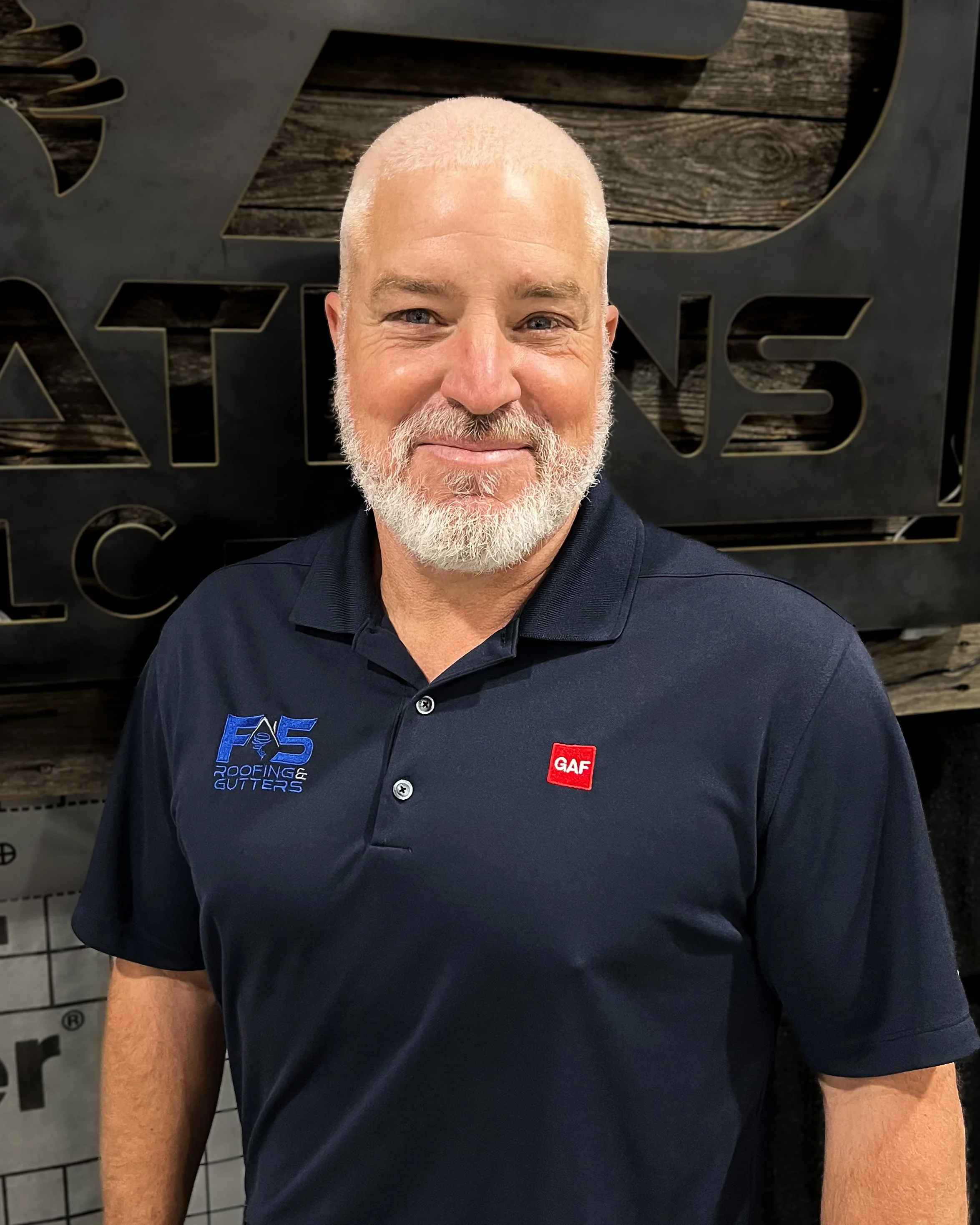 A smiling man with a shaved head and a beard is posing for a selfie in front of a wall with decorative panels. He is wearing a dark polo shirt with logos on the chest.