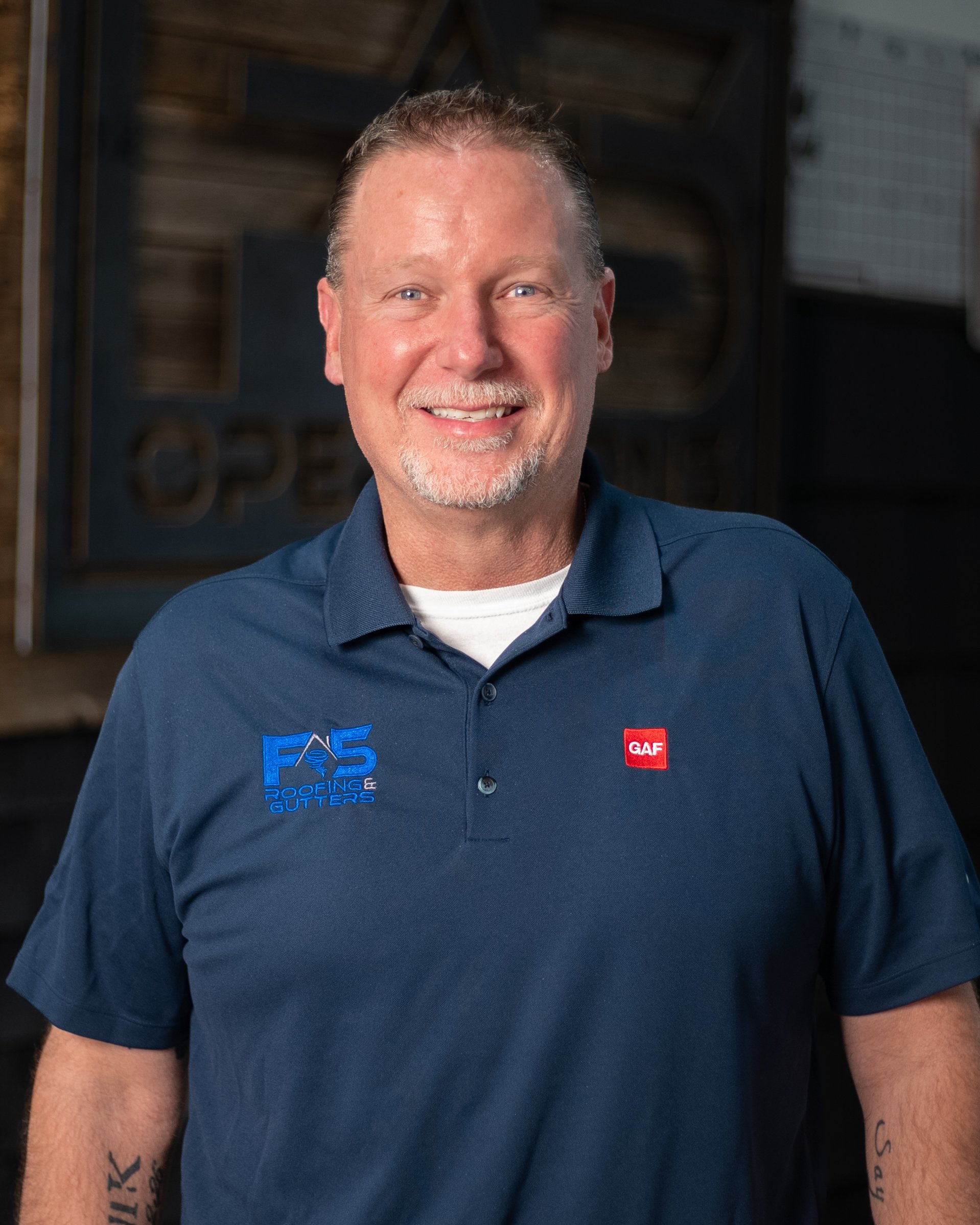 A smiling man with short brown hair and a beard, wearing a blue polo shirt with logos for F5 Roofing Gutters, Under Armour, and Atlas, standing outdoors in front of a glass window.