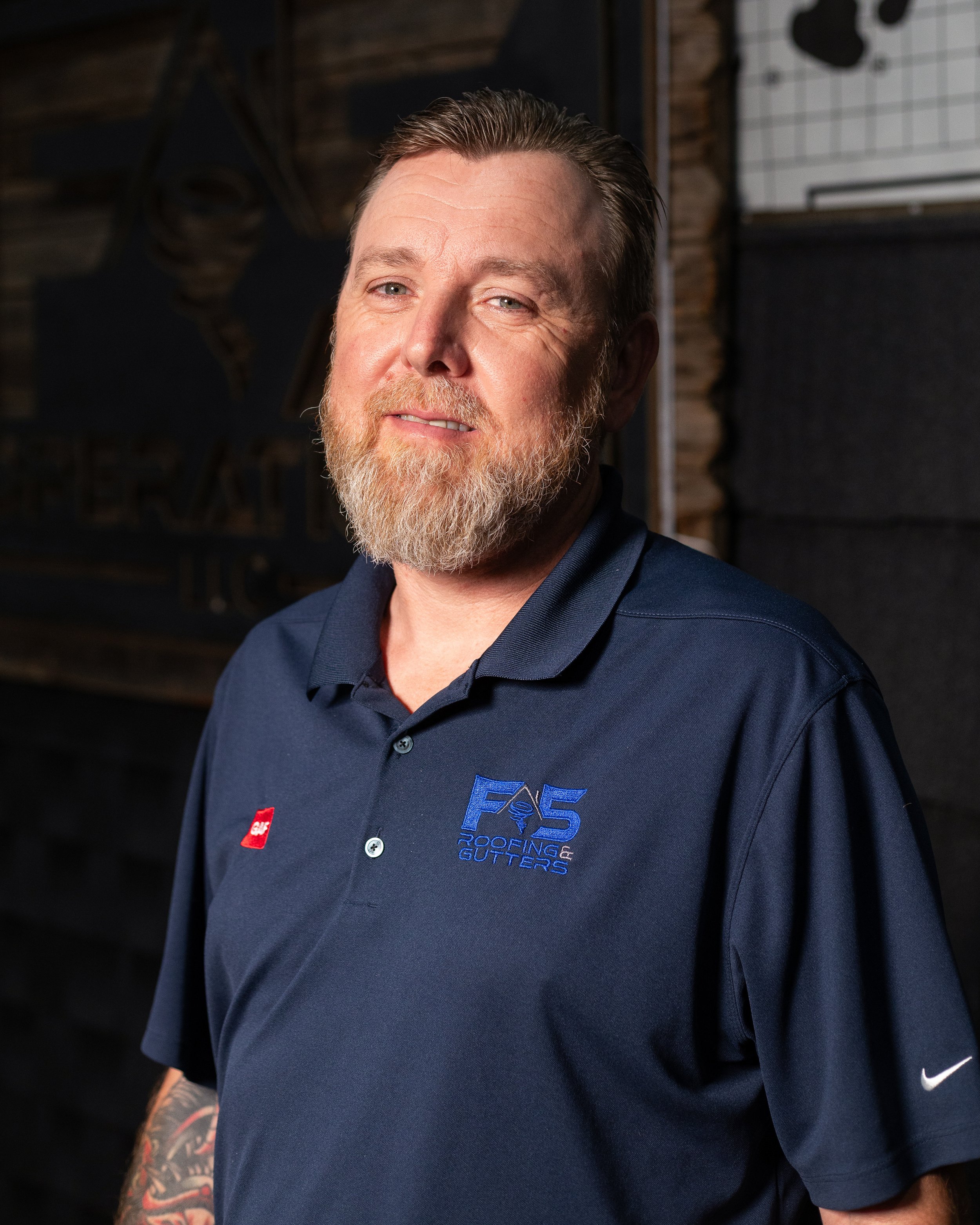 Close-up of a man with light skin, reddish-brown beard, and slicked-back hair, wearing a navy blue polo shirt with logos for roofing and gutter services, standing indoors against a wooden wall with framed artwork and a whiteboard visible in the background.