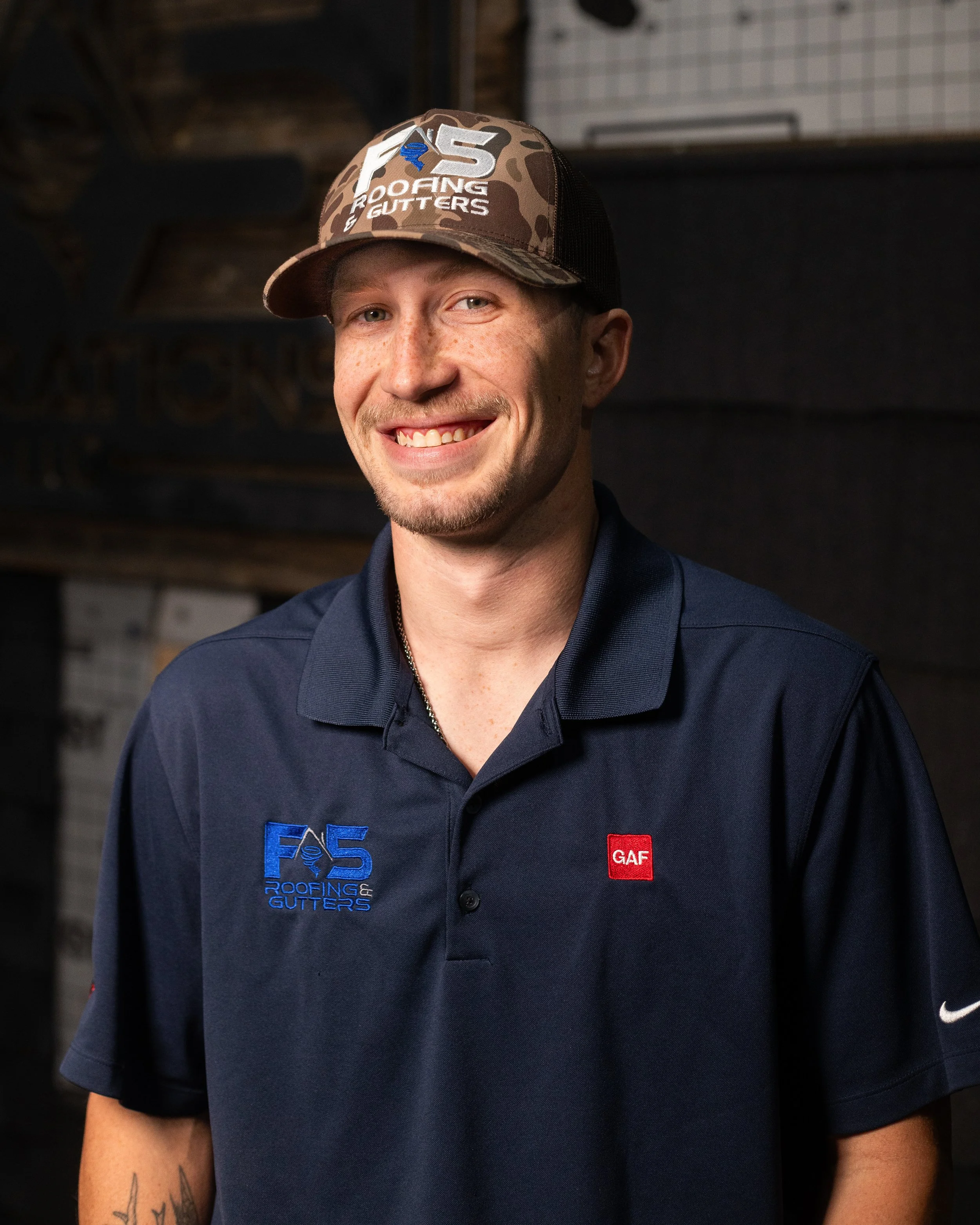 A man smiling, wearing a baseball cap and a navy blue shirt with logos for a roofing and gutters company, standing in front of a background with dark wooden and brick elements.