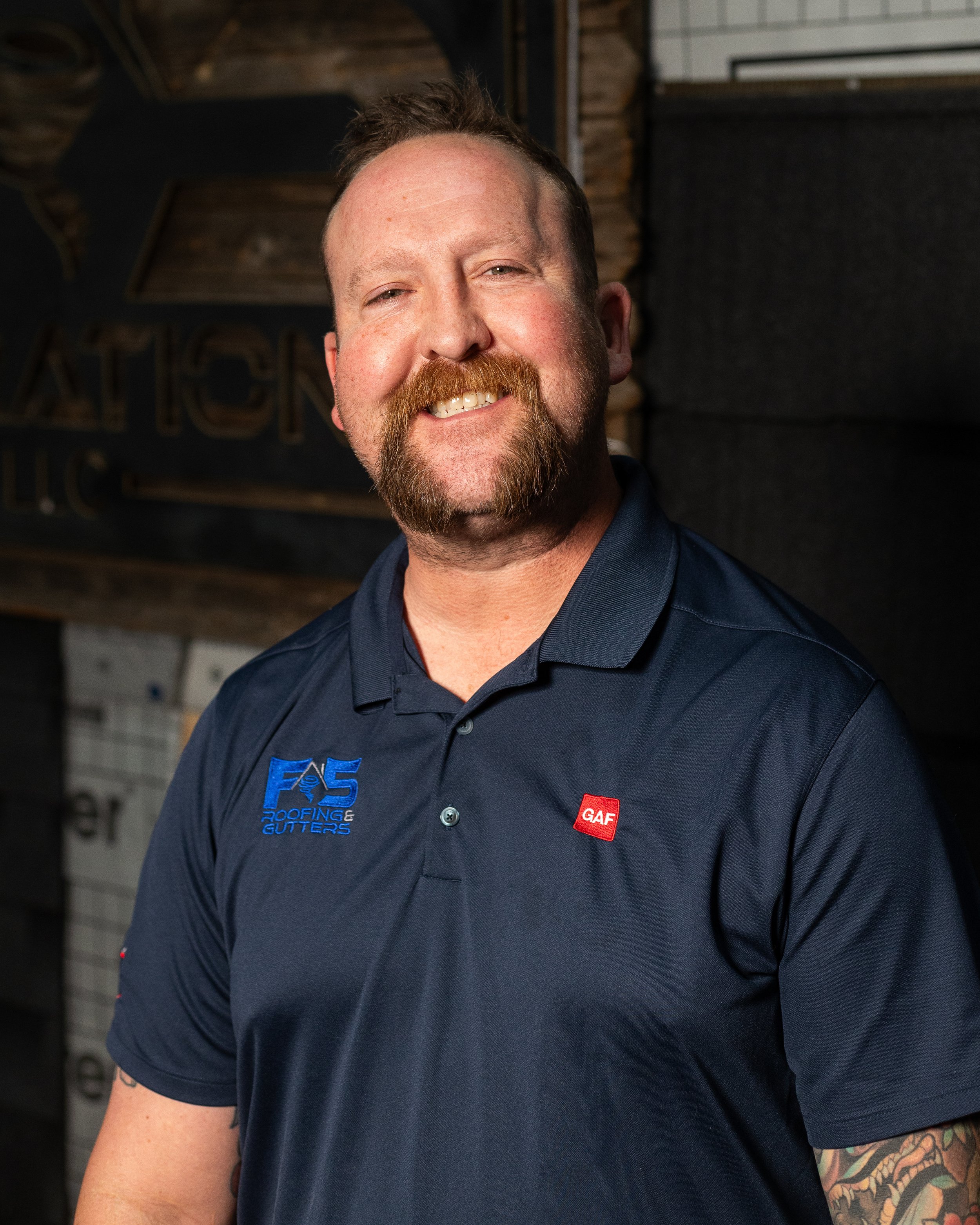 A smiling man with a beard and tattoos wearing a navy blue polo shirt with the logos 'FAS Roofing & Gutters' and 'GAF' standing in front of a wall with siding and acoustic panels.
