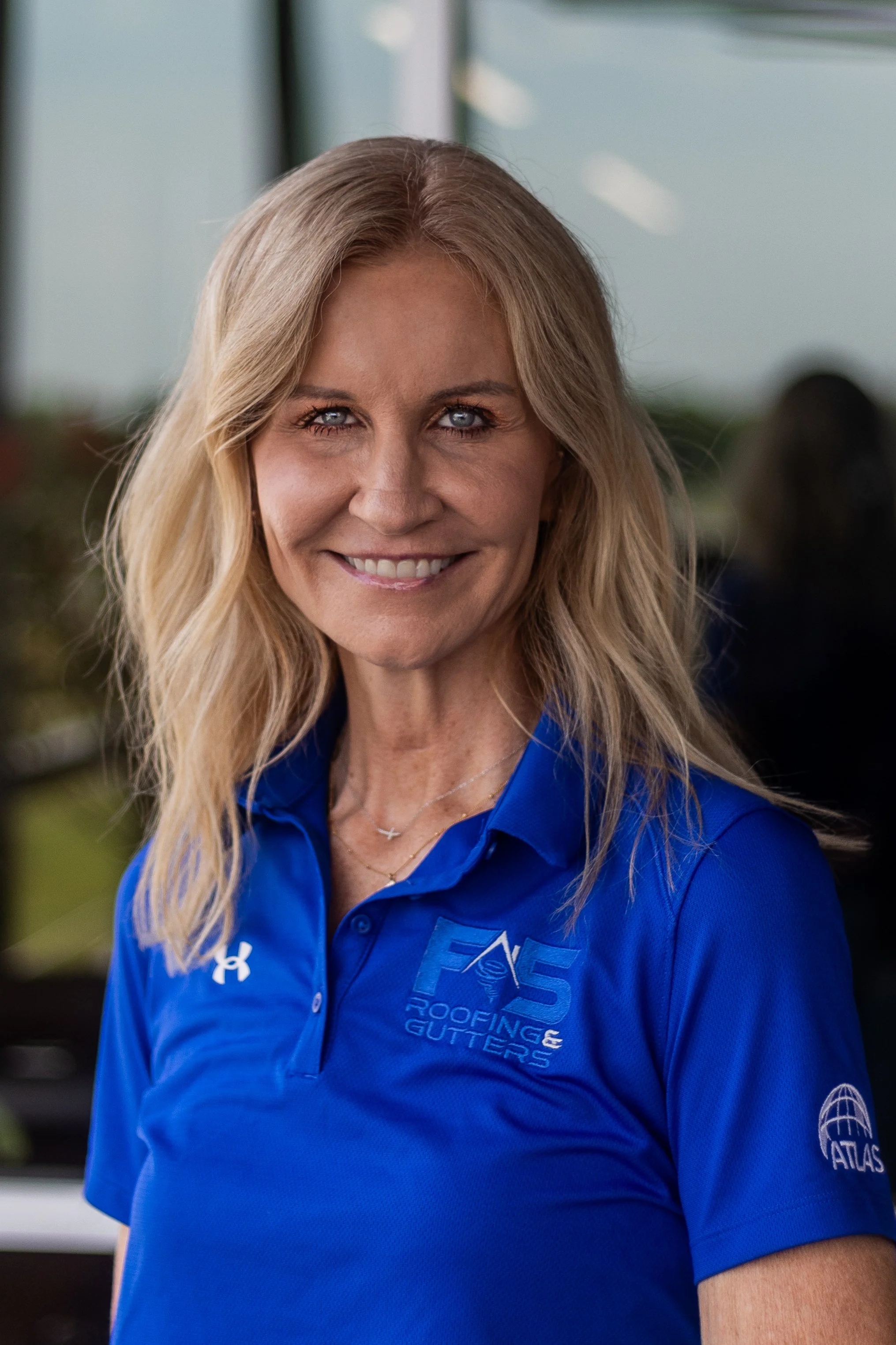 A smiling woman with blonde hair wearing a blue polo shirt with company logos, standing outdoors.