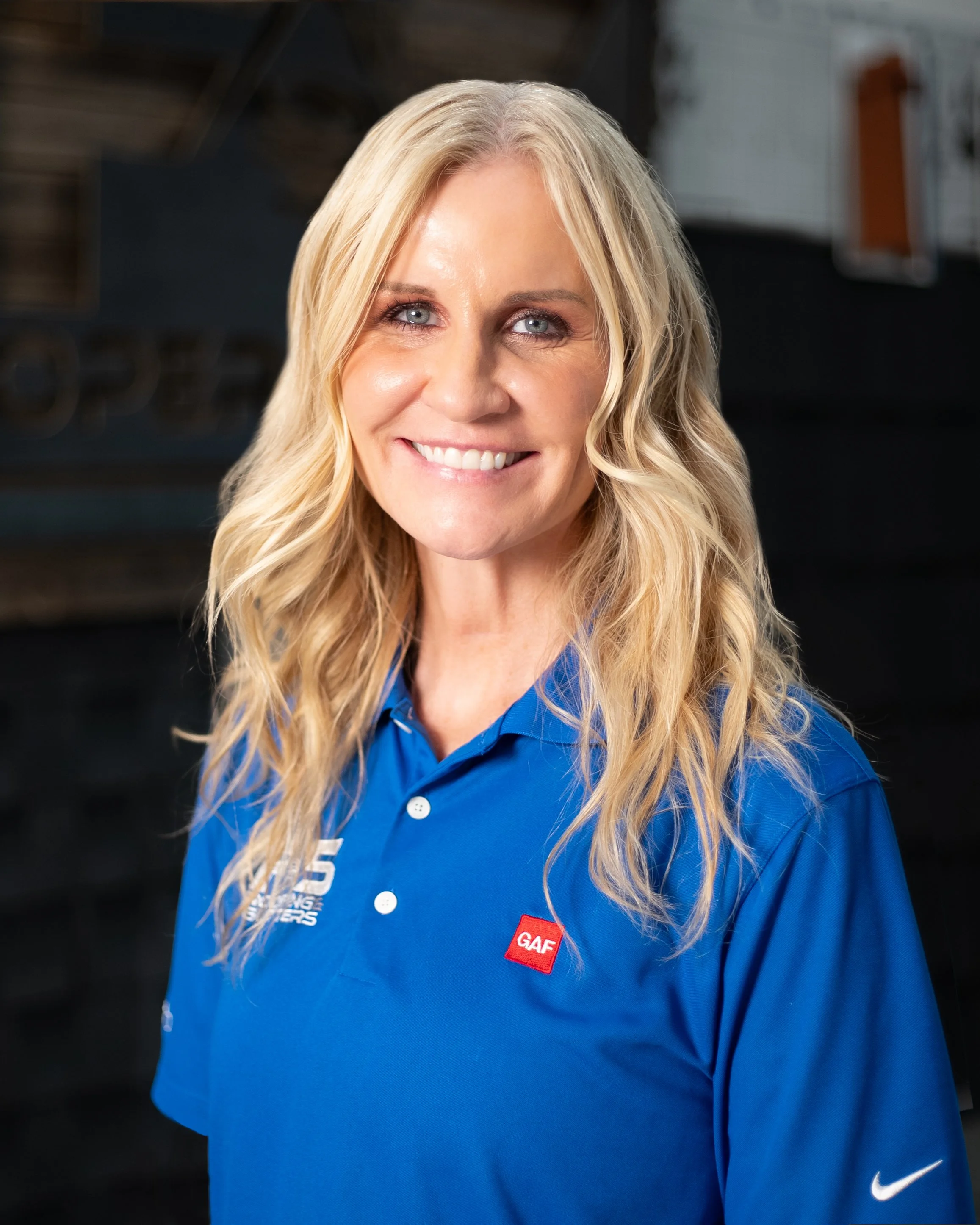 A smiling woman with blonde hair wearing a blue polo shirt with company logos, standing outdoors.