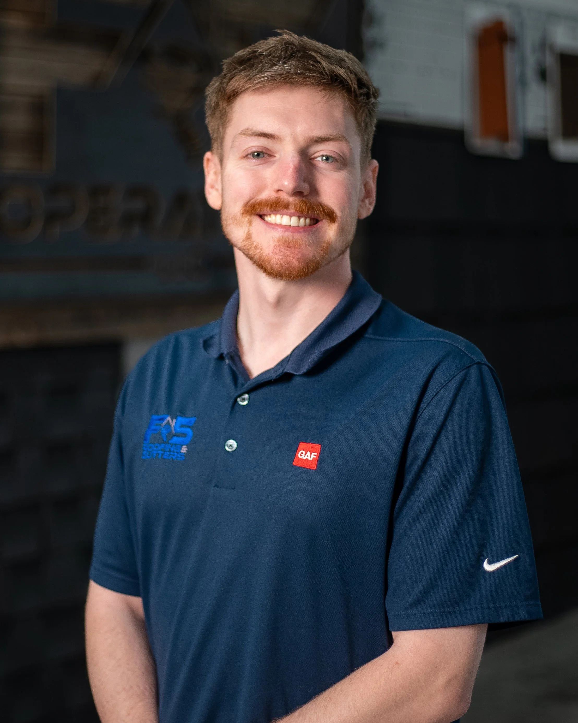 A smiling man in a blue polo shirt with a roofing logo, standing outdoors.