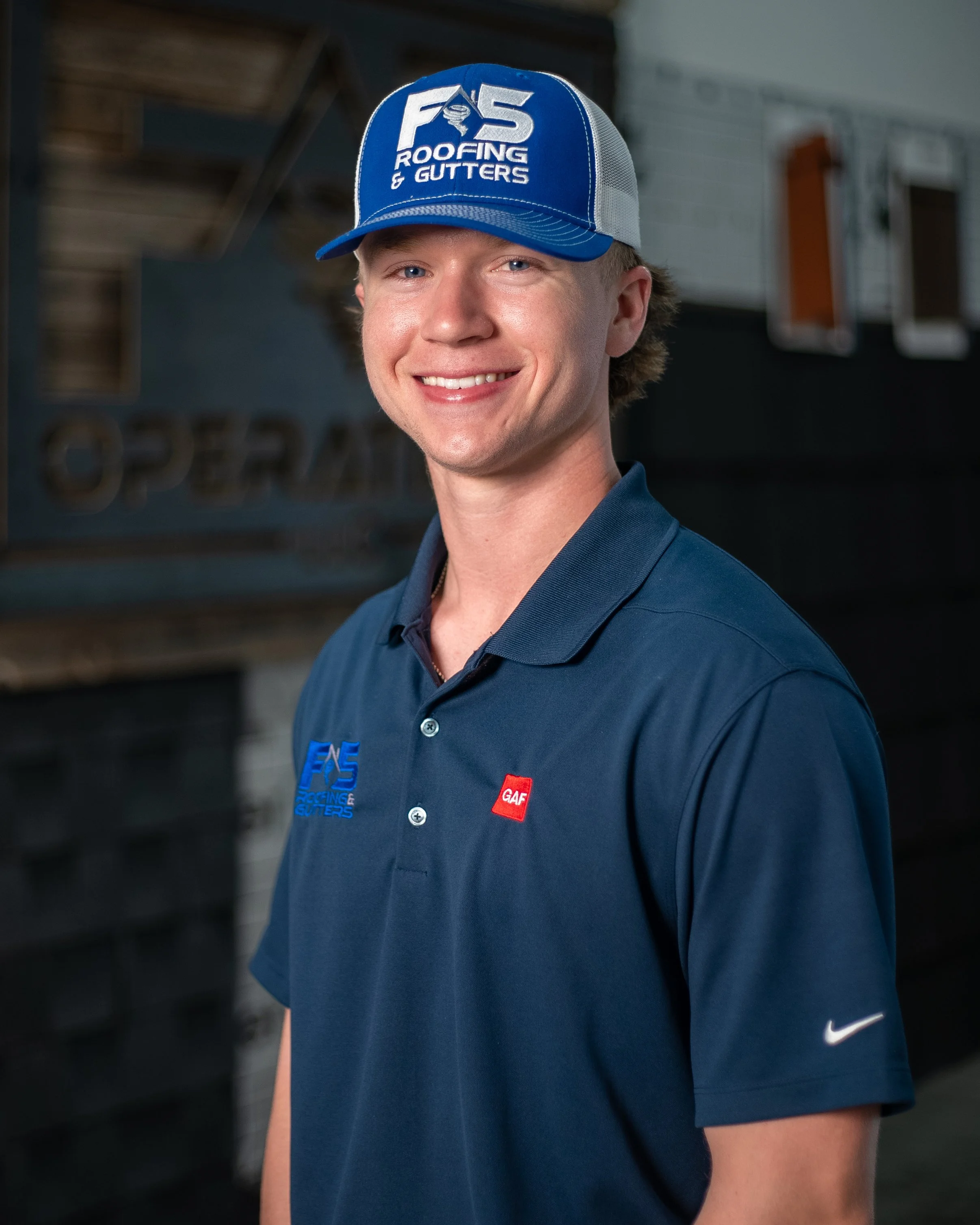 Young man smiling into camera with a blue ball cap that reads "F5 Roofing and Gutters" and wearing a navy blue polo