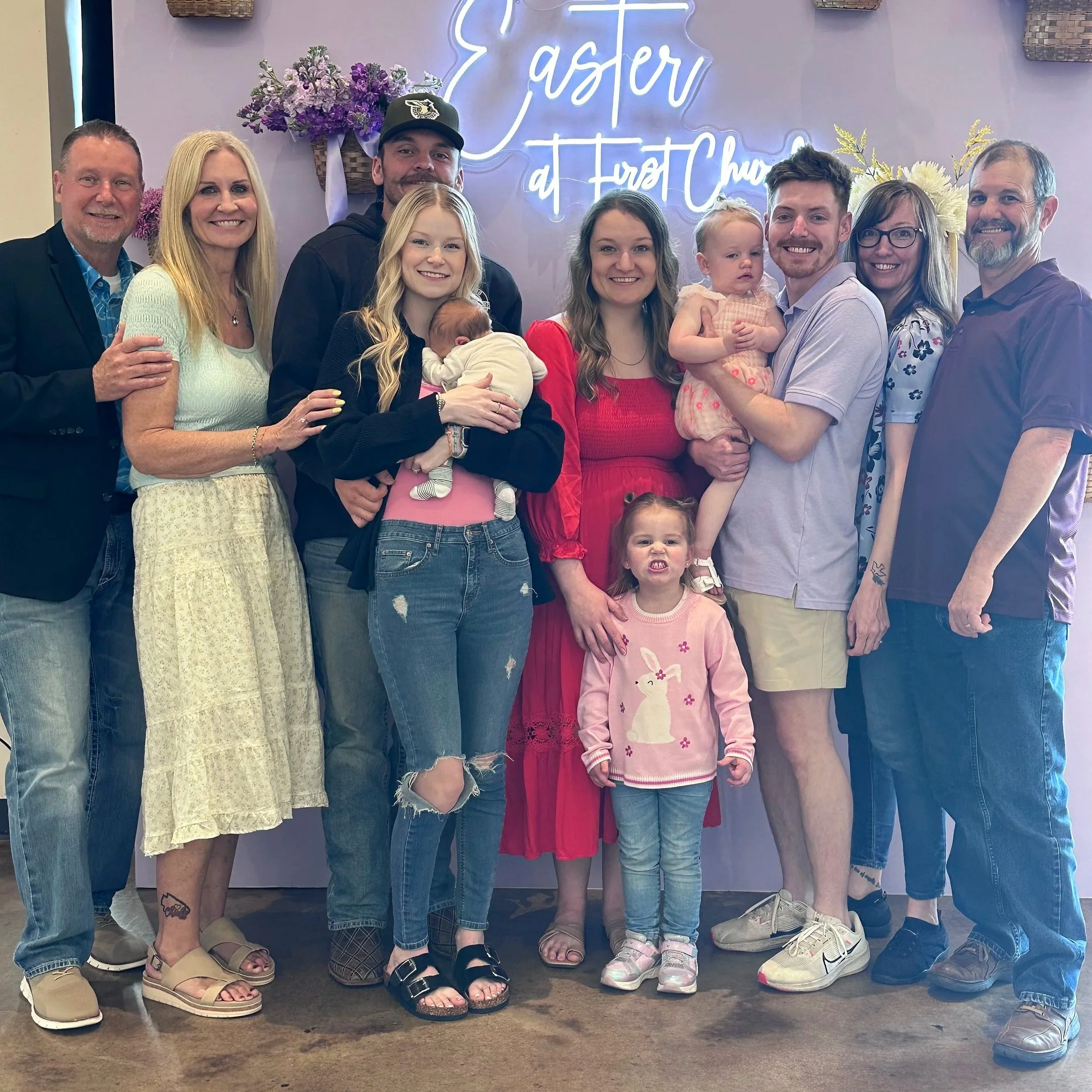 A group of ten people, including children, standing in front of a pastel pink wall with a neon sign that says 'Easter at First Church.' They are smiling and dressed casually, with some holding small children or babies, and there are flower arrangements on the wall behind them.