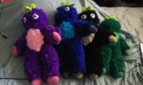 Four colorful plush toy dolls with big eyes, pink, blue, black, and green in color.