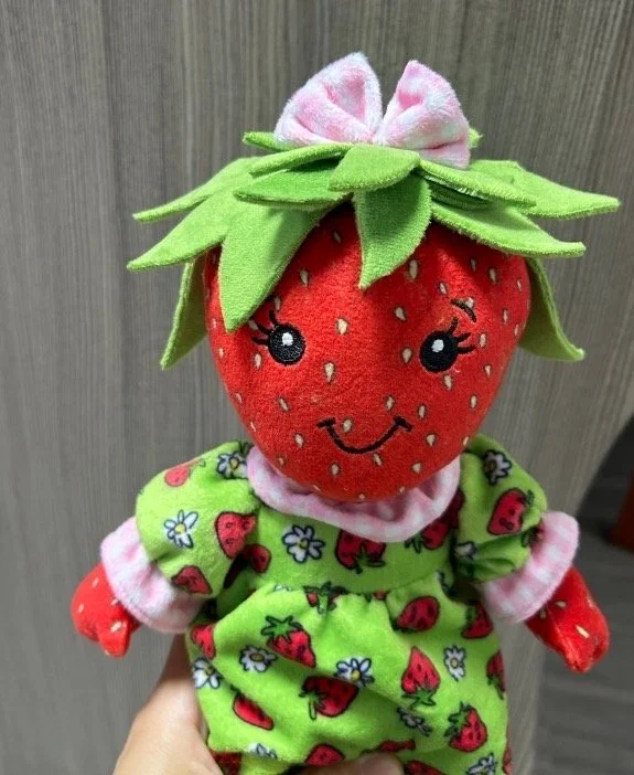 A plush strawberry doll with an embroidered smiling face, dressed in a green strawberry-patterned outfit with pink trim, topped with green leaves and a pink bow on her head.