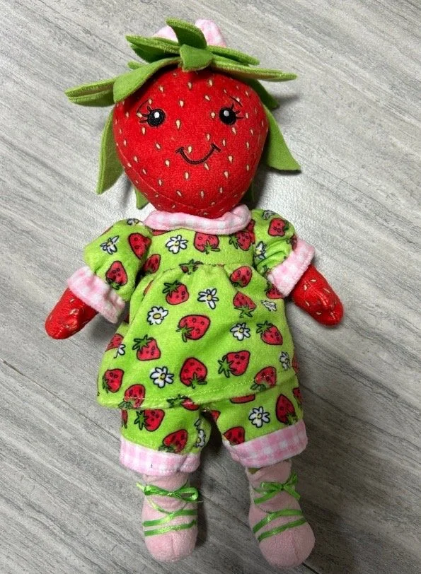 A strawberry plush toy with a smiling face, dressed in a green strawberry-patterned outfit, pink and white striped pants, and pink shoes with green laces.