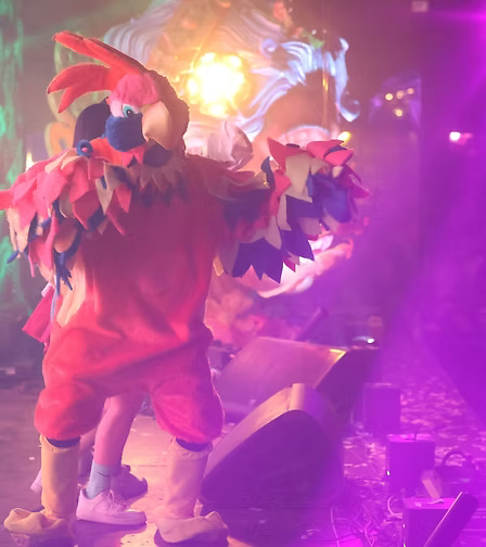 Personaggio in costume da uccello colorato con piume rosse e blu, in un ambiente festoso con luci colorate.