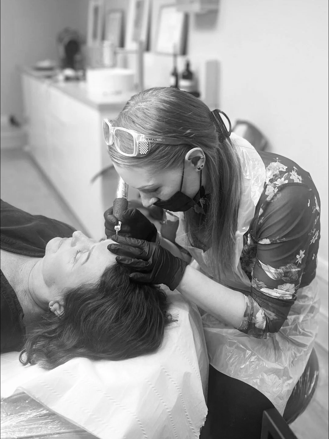 A permanent makeup artist carefully applies microblading machine hairstrokes to a clients eyebrows in a brightly lit salon