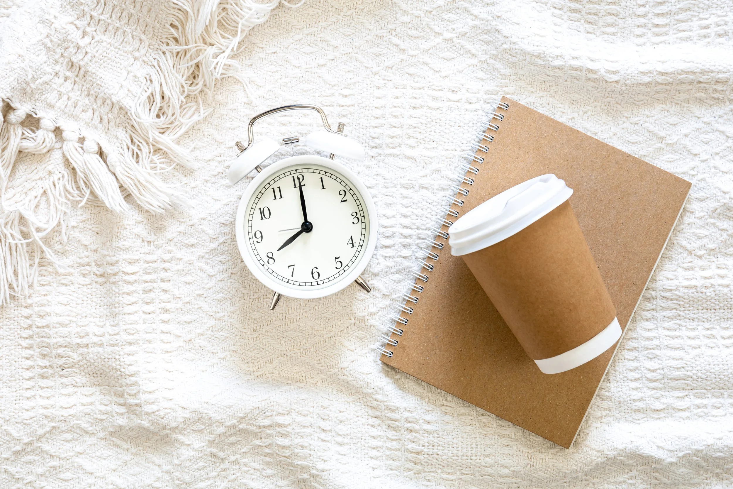 A neutral bed cover, alarm clock, and takeaway coffee cup illustrating a relaxing morning routine before a permanent makeup appointment at Salon 21 Bristol