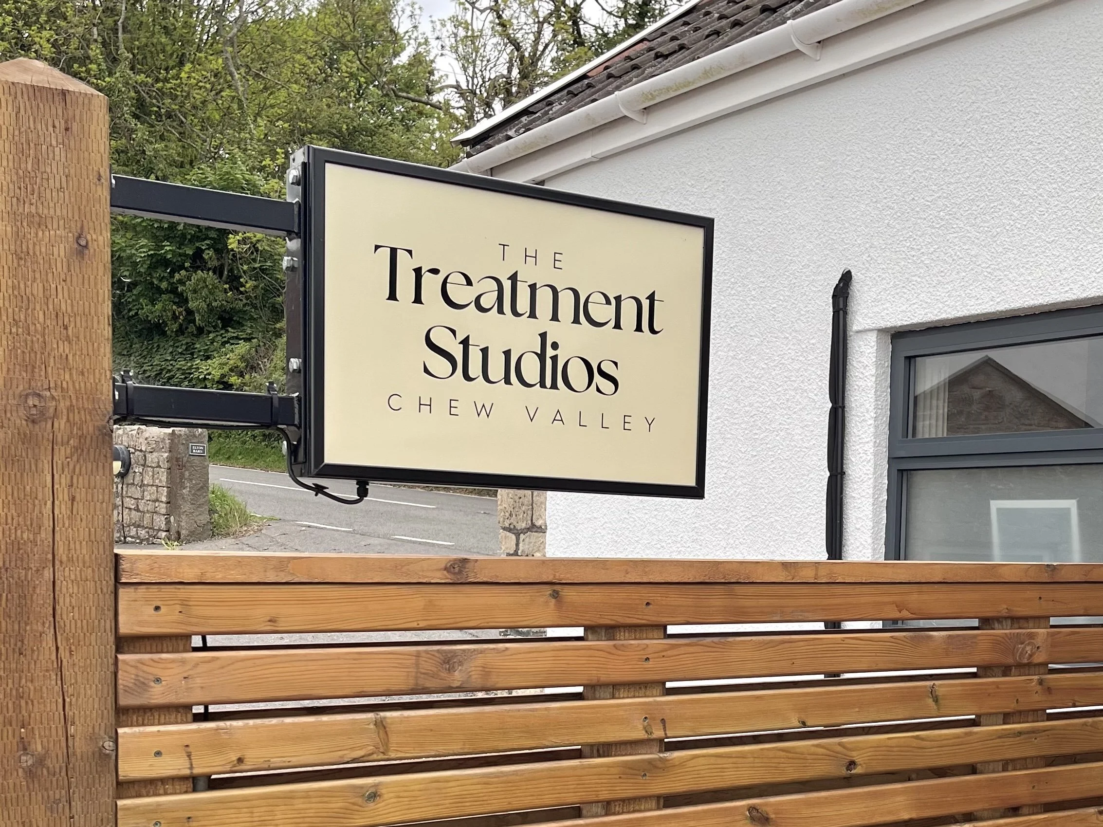 The front entrance of the salon featuring a black black and beige modern sign that reads 'The Treatment Studios' Chew Valley on a modern wooden fence