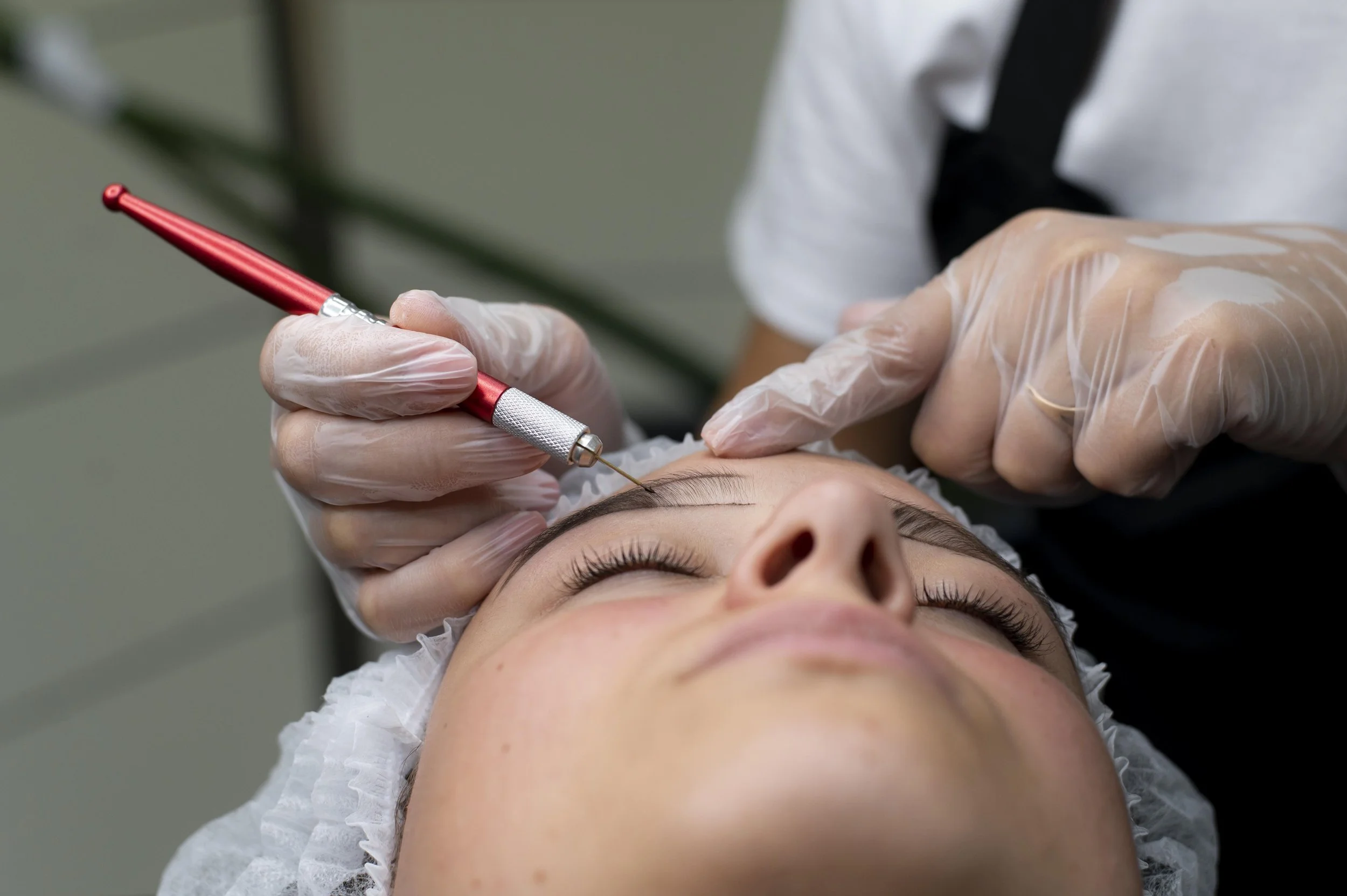 permanent makeup artist wearing gloves performing precise microblading strokes on a client's eyebrows in a clean studio