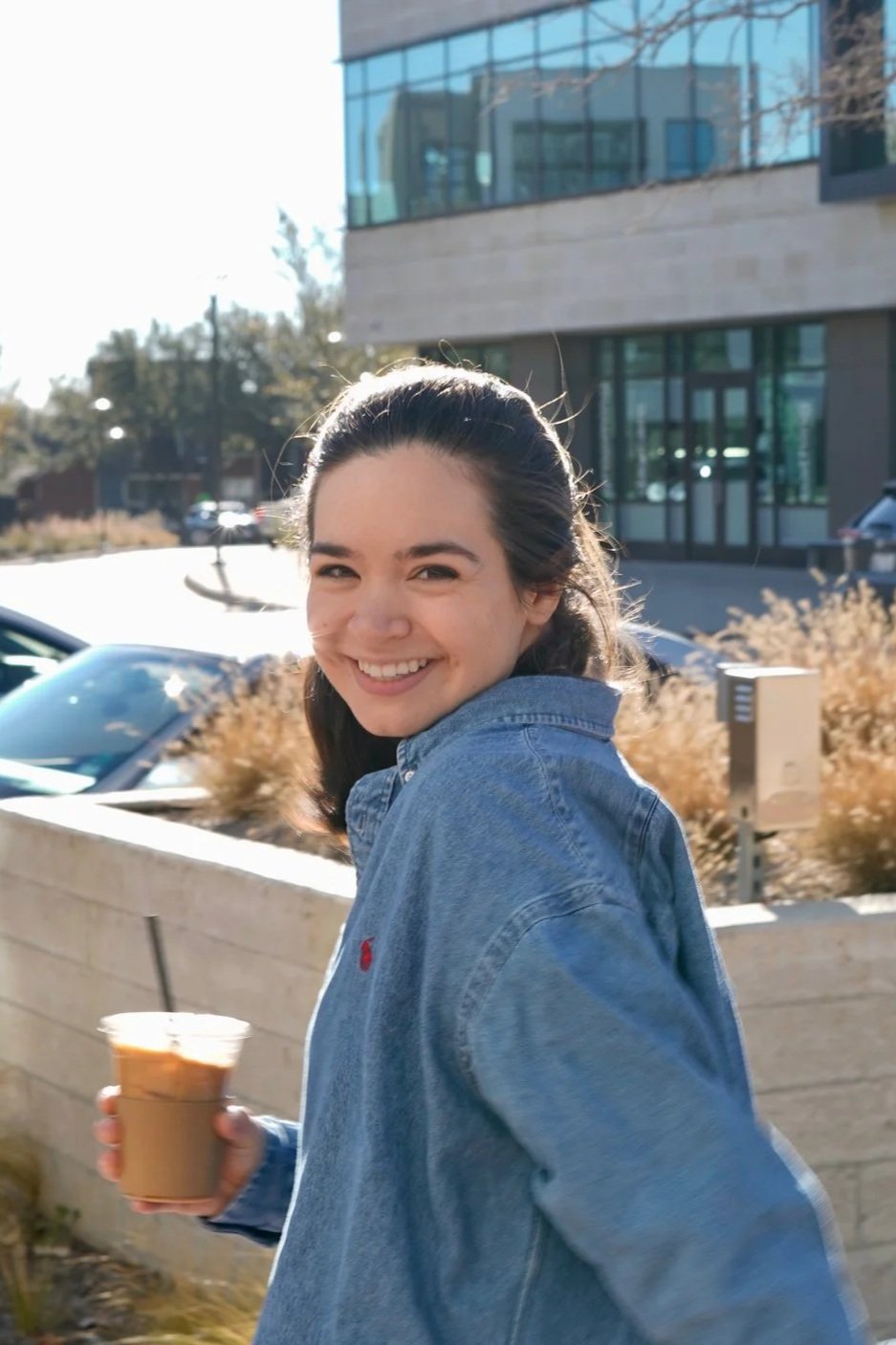 A young woman with dark hair, wearing a denim jacket, smiling while holding an iced coffee outside on a sunny day, in front of a modern building with large glass windows.