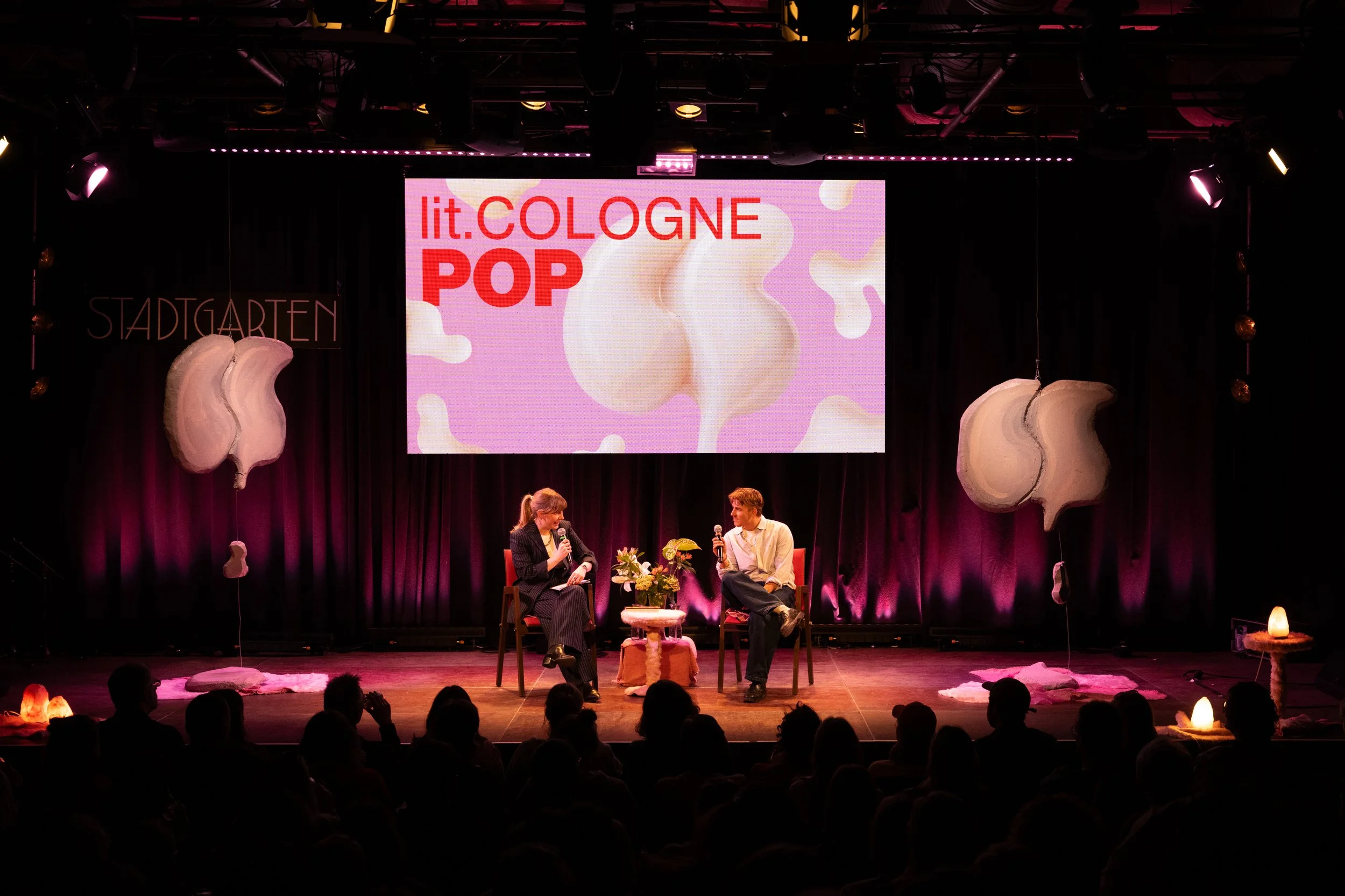 Stage at lit.Cologne POP event with two speakers, a woman and a man, sitting and talking into microphones. Large screen behind them displays 'lit.COLOGNE POP' and a pink abstract background. Decorations include large plush clouds and soft glowing lights.