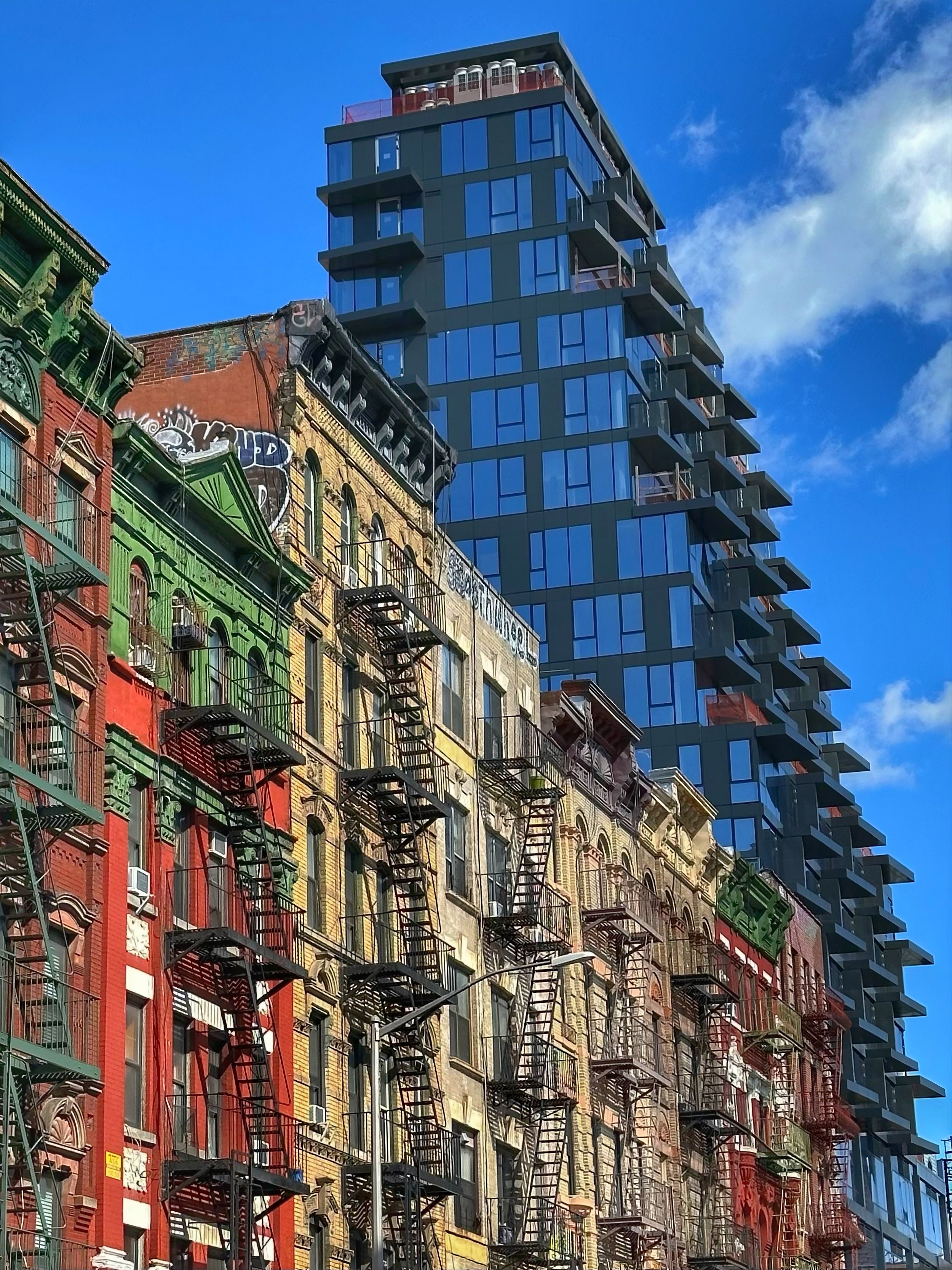 TENEMENTS AND TOWERS -- New York, NY (March 14, 2026) -- Old tenements lining Henry Street in NYC's Chinatown and the Lower East Side have been joined by a new residential tower looming above. The stepped balconies of the new tower resonate with the 