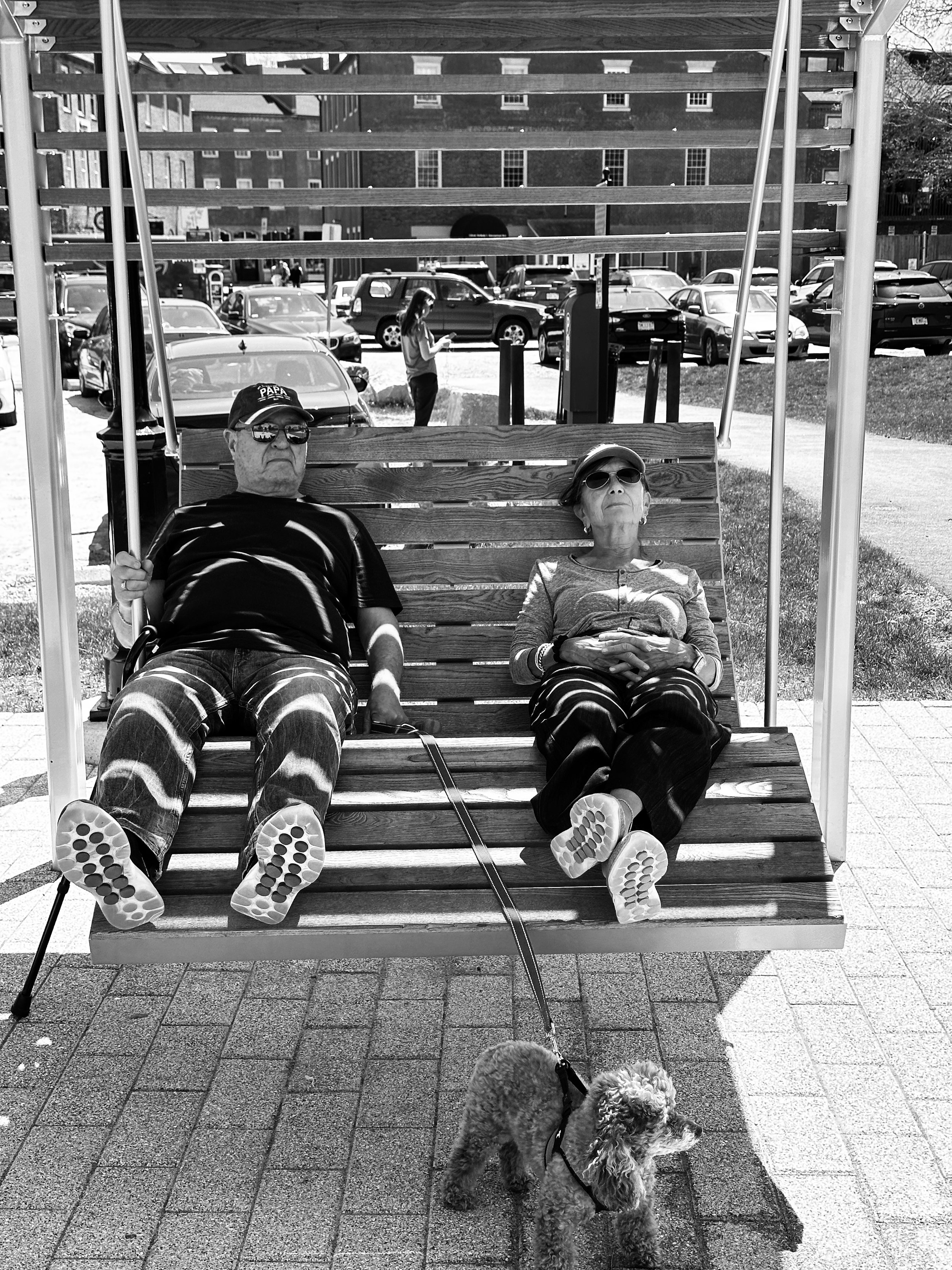 STRIPED BY THE SUN - Newburyport, Massachusetts (May 5, 2026) - A couple relaxes on the swinging benches at Newburyport's central waterfront on an unseasonably warm spring day. This ordinary moment is amplified by the striping of sunlight on their cl
