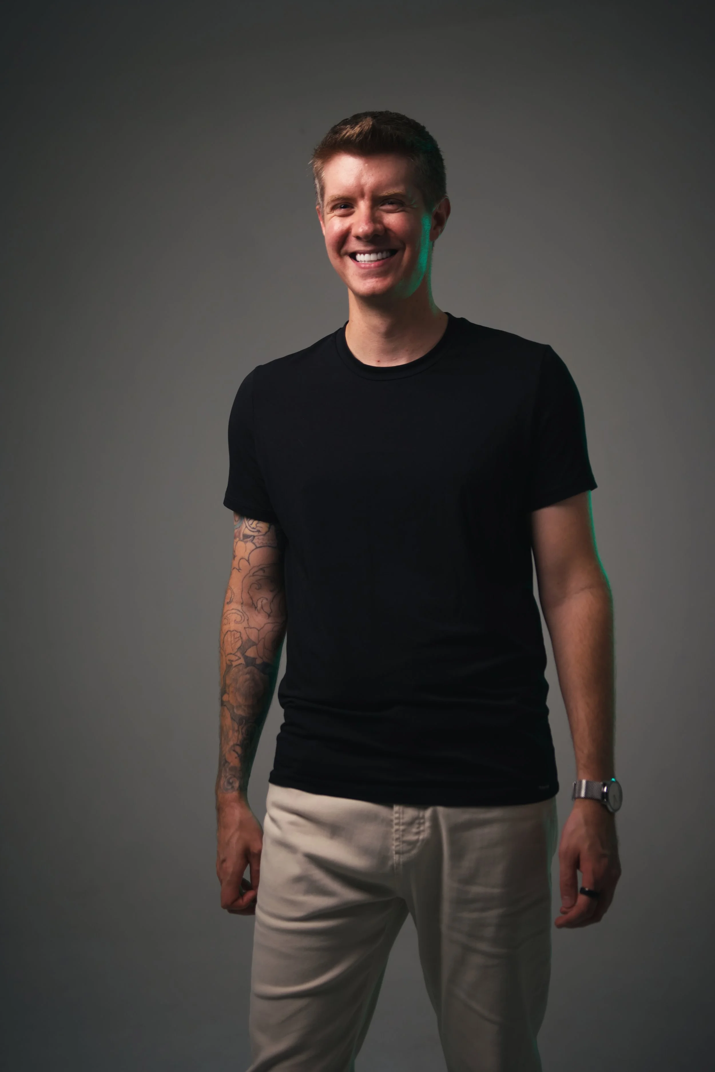 A young man with short brown hair, smiling, wearing a black t-shirt, beige pants, a wristwatch, and a ring, standing against a gray background.