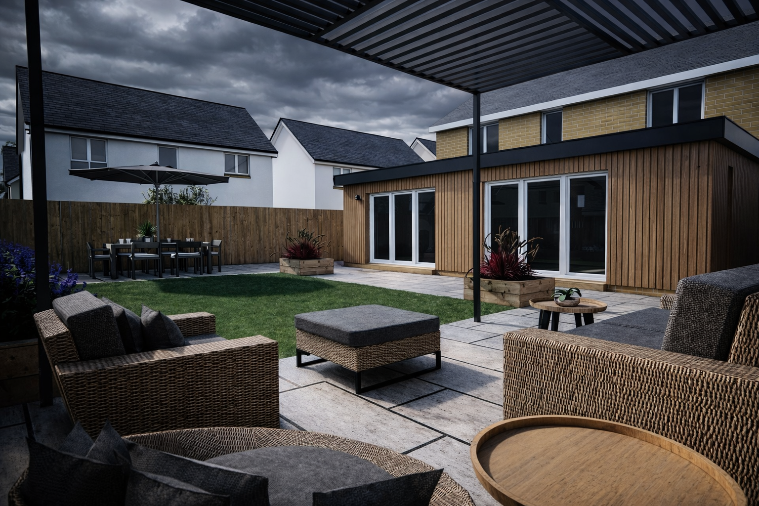 A modern backyard with a covered patio, wicker furniture, a small table, green lawn, potted plants, and a nearby extended patio area with dining table and chairs under an umbrella. Modern houses with varied facades are visible in the background under a cloudy sky.