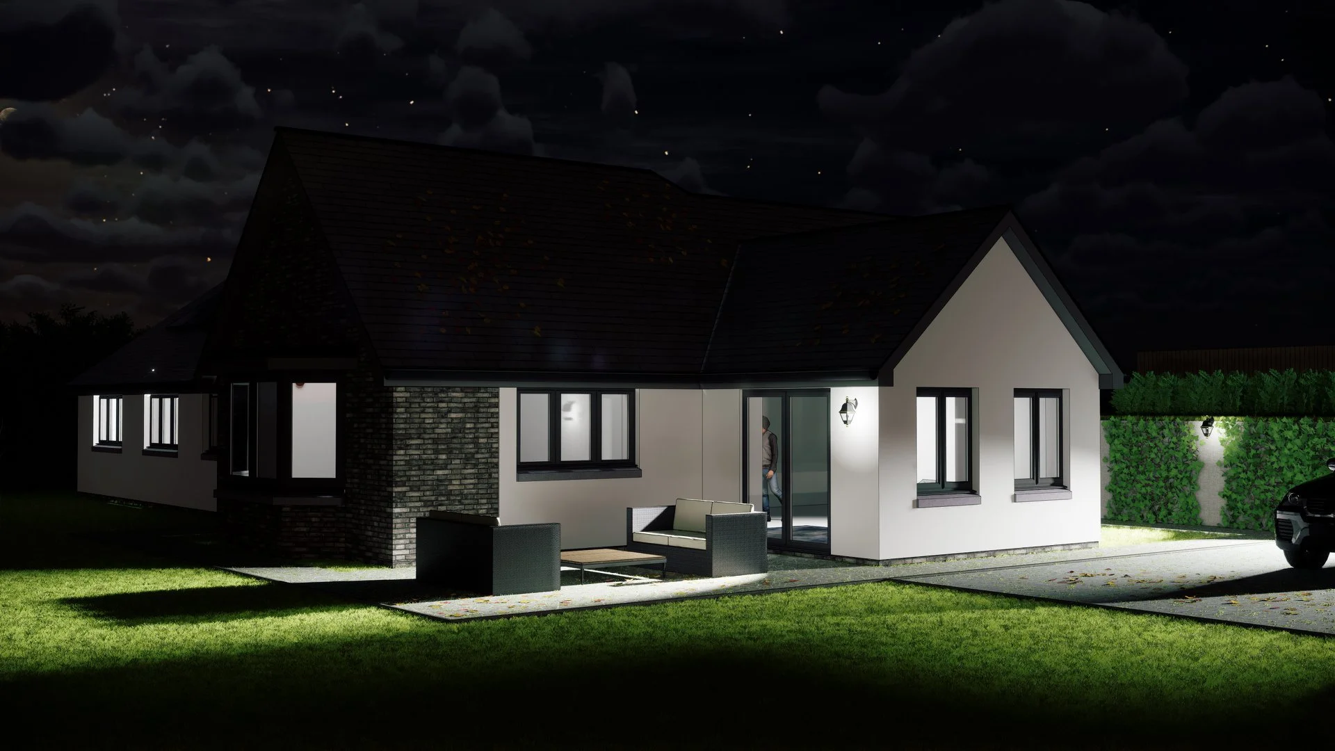 Nighttime view of a modern single-story house with lit windows, outdoor seating area, and a person standing near the door. The house has a gabled roof, white exterior walls, and a brick section. There is a driveway with a parked car and a lush green lawn.