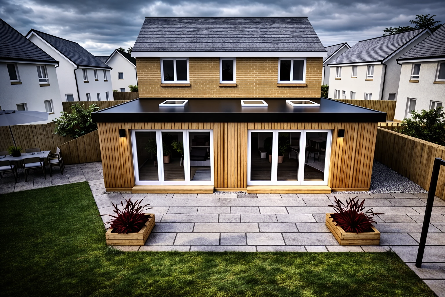 Modern backyard with a small wooden house featuring large glass sliding doors, potted plants inside, and a black flat roof with skylights. The yard has a paved patio, a green lawn, planters with red plants, outdoor dining table with chairs, and a wooden fence surrounding the area under a cloudy sky.