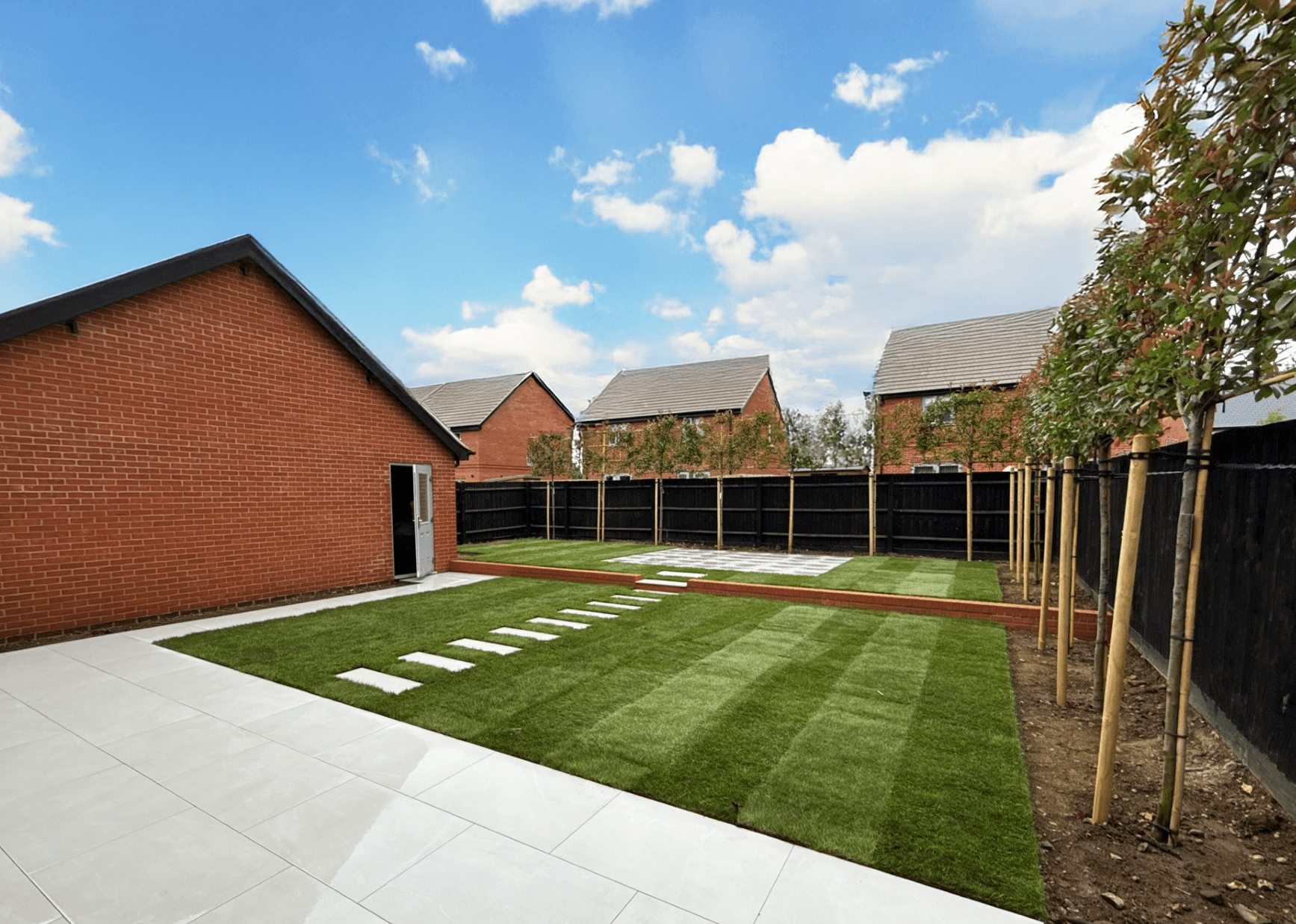Backyard with a brick shed, a black fence, green lawn, stepping stones, and newly planted trees under a partly cloudy sky.