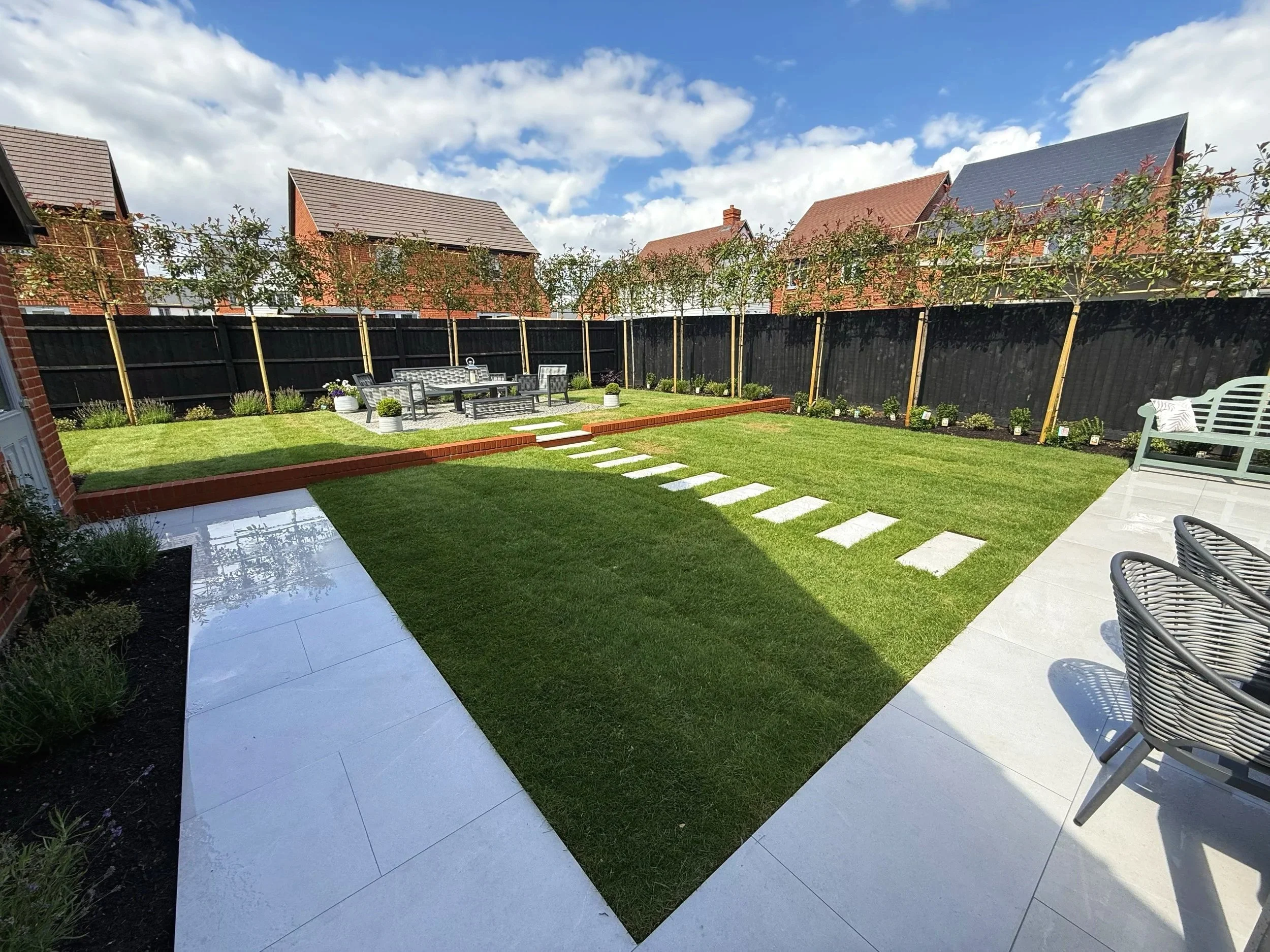 A well-maintained backyard garden with a lush green lawn, stepping stones, outdoor furniture, potted plants, and a black wooden fence, with houses and blue sky with clouds in the background.