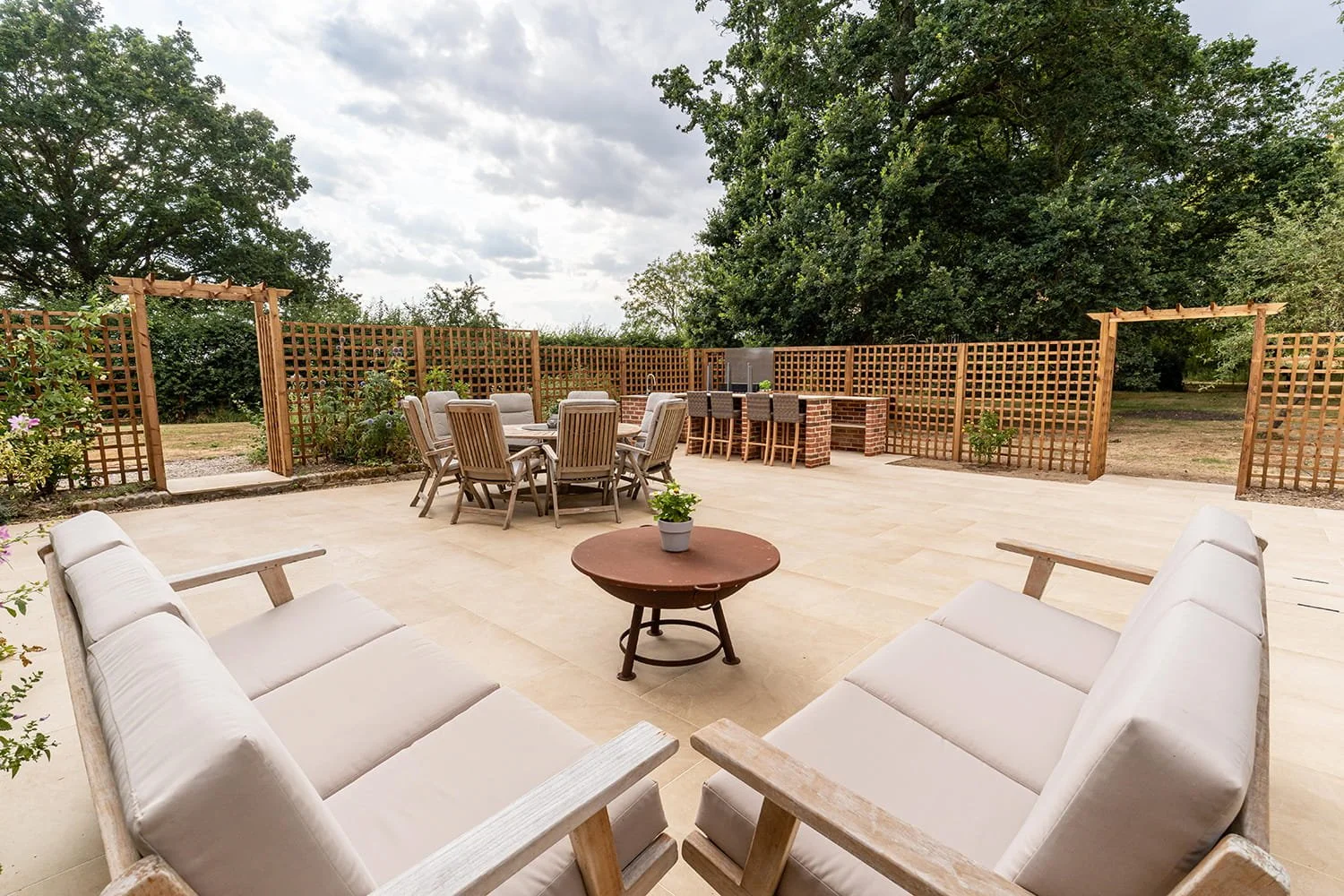 Outdoor patio with outdoor furniture including two cushioned chairs, a round coffee table with a potted plant, a rectangular dining table with eight wooden chairs, a bar area with four barstools, and a wooden lattice fence with plants, surrounded by trees under cloudy sky.