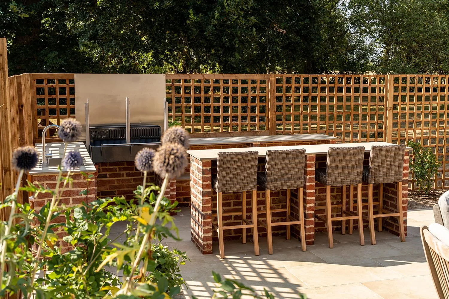 Outdoor patio area with brick bar counter, four wicker bar stools, a grill, and a wooden privacy fence, surrounded by greenery.
