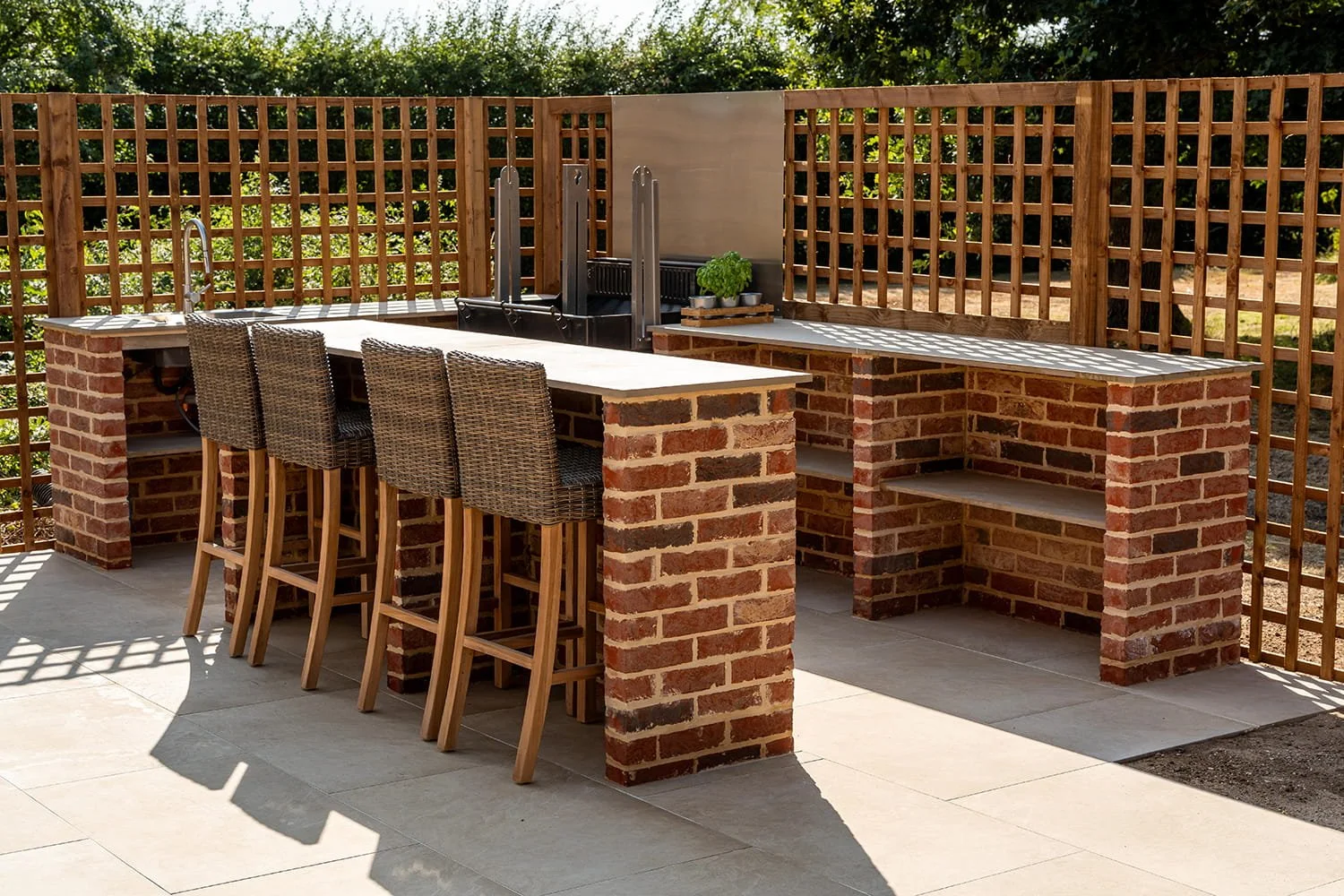 Outdoor kitchen with brick countertops, bar seating with four wicker chairs, a sink, and a fenced background with greenery.