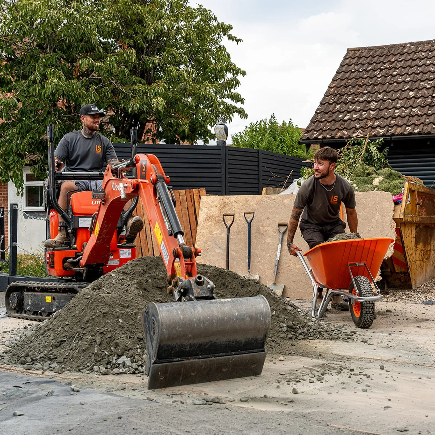 Two construction workers working on a street repair project. One is on a mini excavator, and the other is pushing a wheelbarrow filled with dirt or gravel. There is a pile of dirt in front, and construction tools including shovels and a dump trailer are visible. Behind them are trees, a house with a tiled roof, and a dark fence.