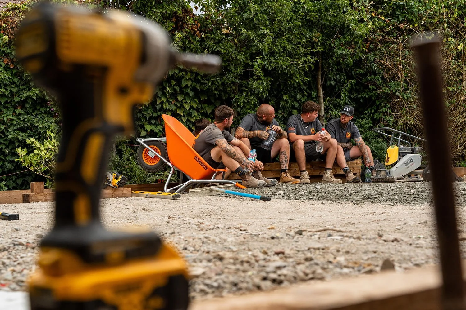Four construction workers sitting on a wooden bench taking a break, with construction tools and equipment around them, and greenery in the background.